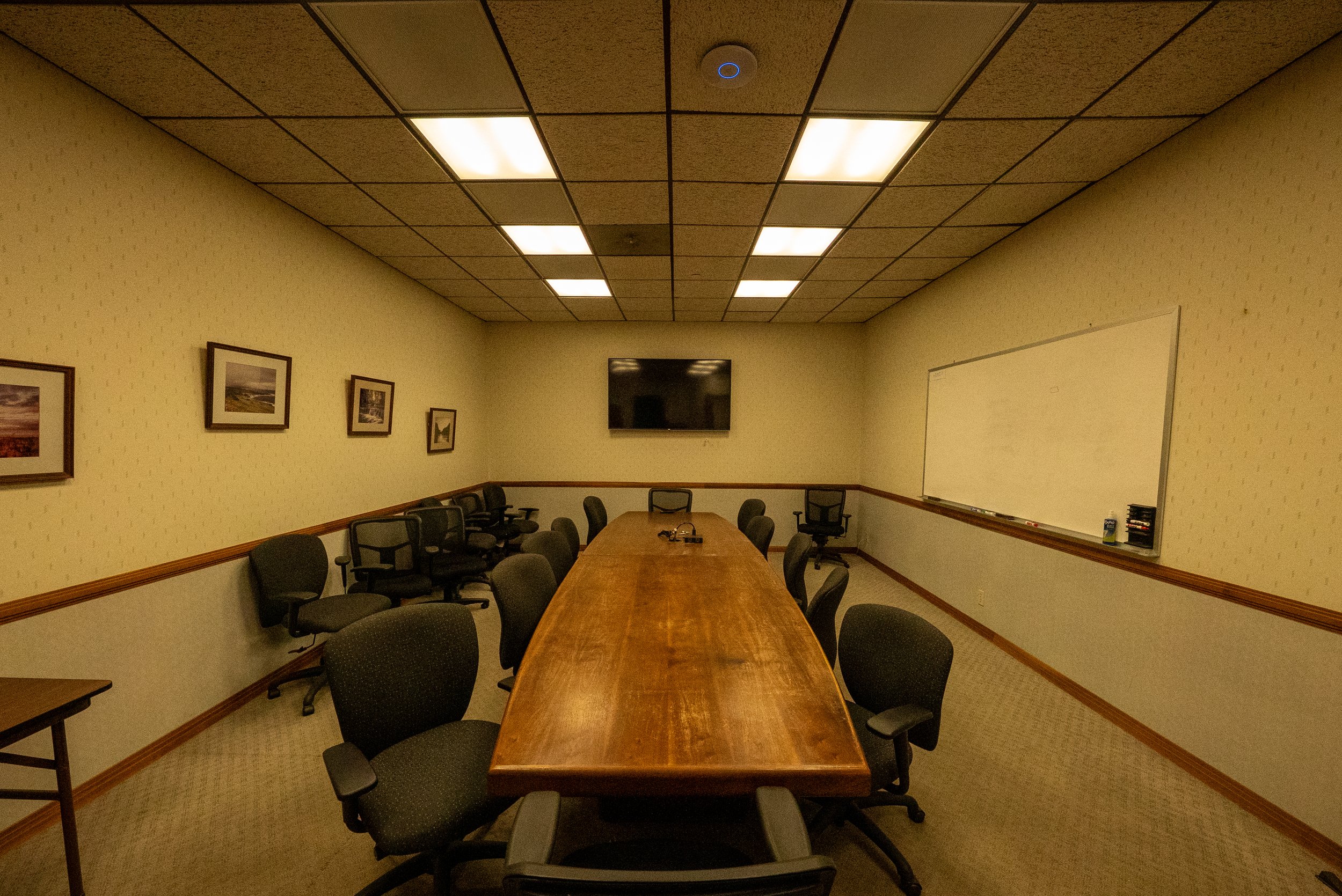 A conference room with a long wooden table, black office chairs, framed pictures on the beige walls, a whiteboard, and a television on the wall under a ceiling with fluorescent lights.