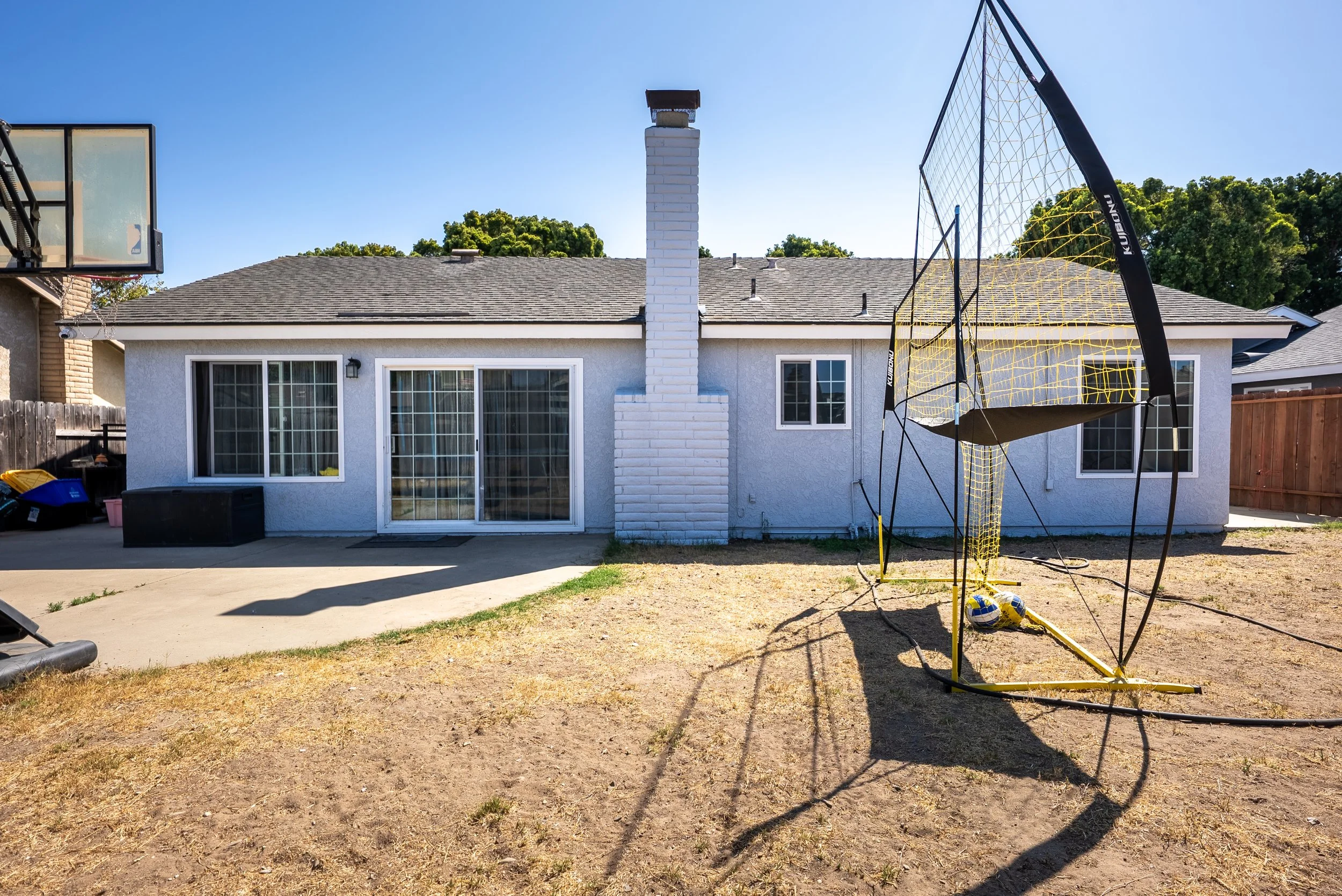 Backyard with a white house, sliding glass doors, a brick chimney, and a trampoline on a dry lawn under a clear blue sky.