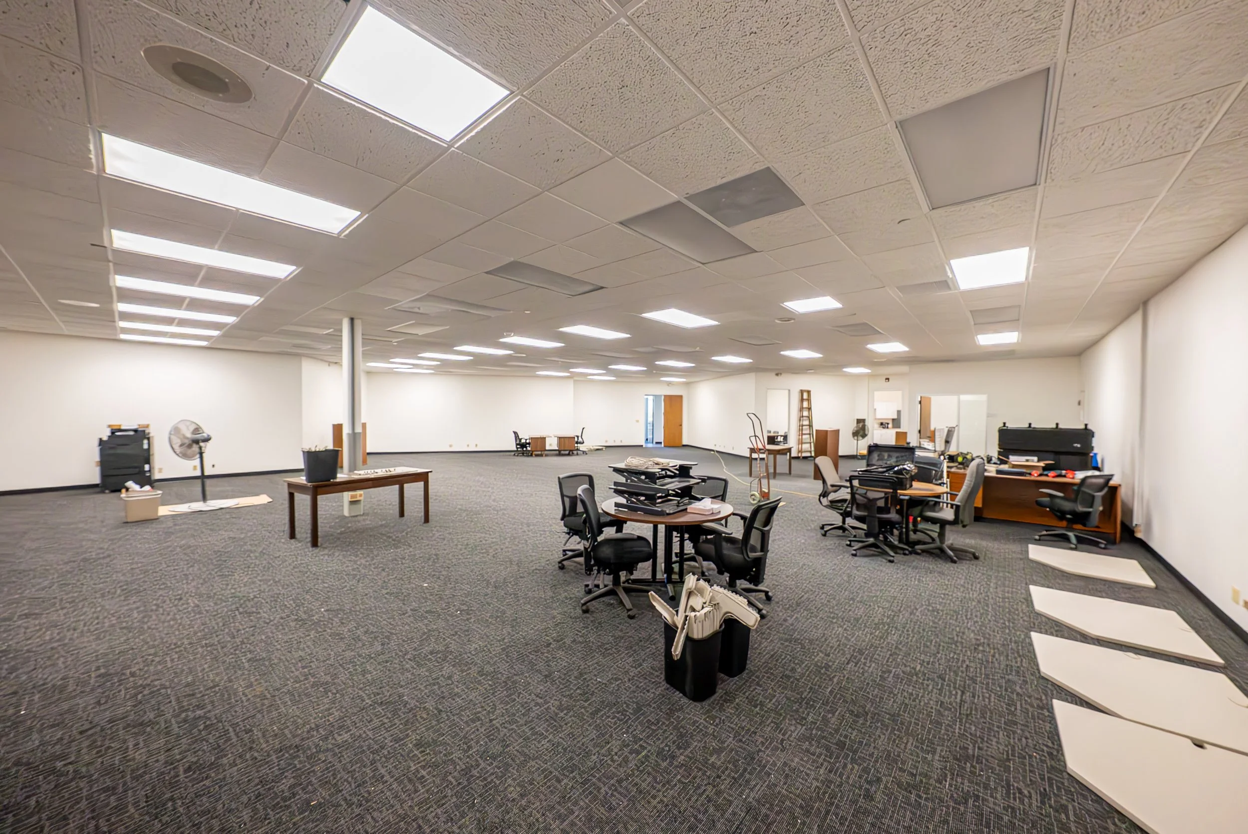 Large, empty office or conference room with scattered office furniture, chairs, tables, and some ceiling tiles removed.