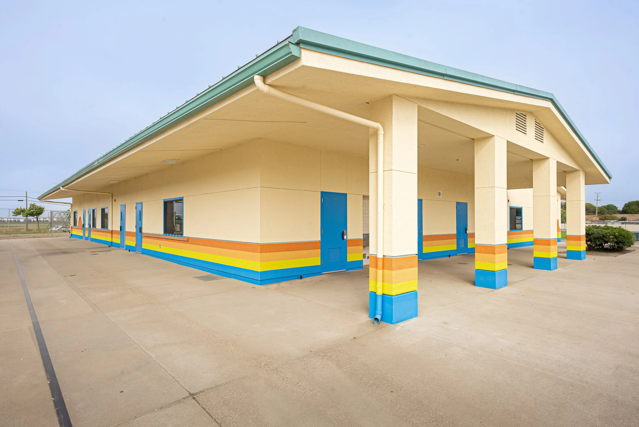 A colorful building with a sloped roof and bright blue doors, located in an outdoor area with a concrete surface and a playground structure in the background.