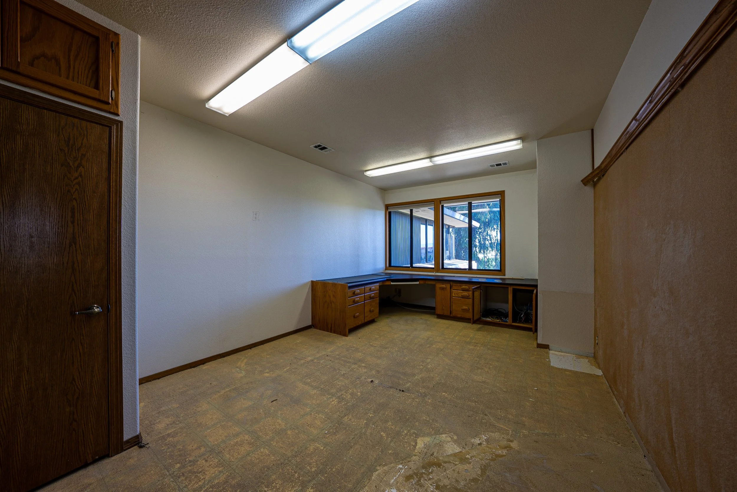 Empty room with wooden cabinets and large window, fluorescent ceiling lights, beige carpet, and a wall with a doorway.