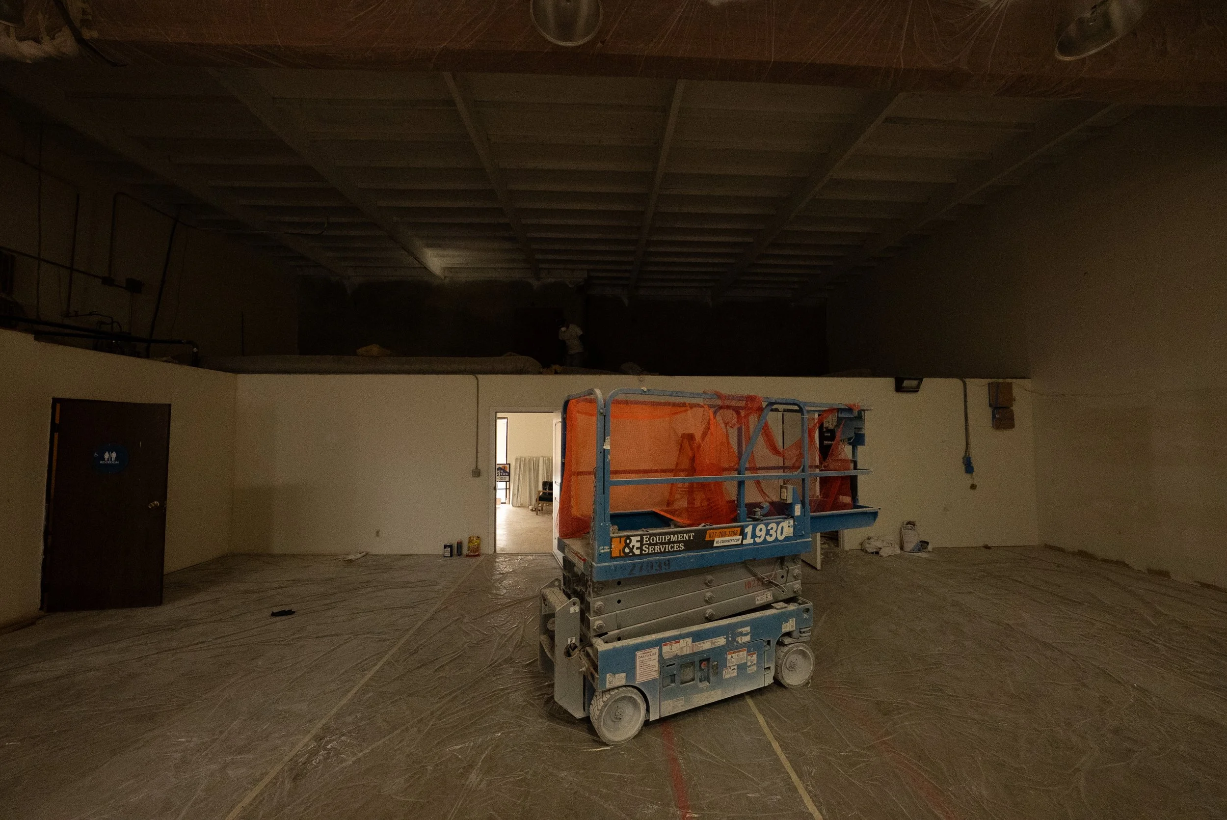 Construction site with a lift and a person in the background, the area is partially finished with a suspended ceiling and construction equipment.