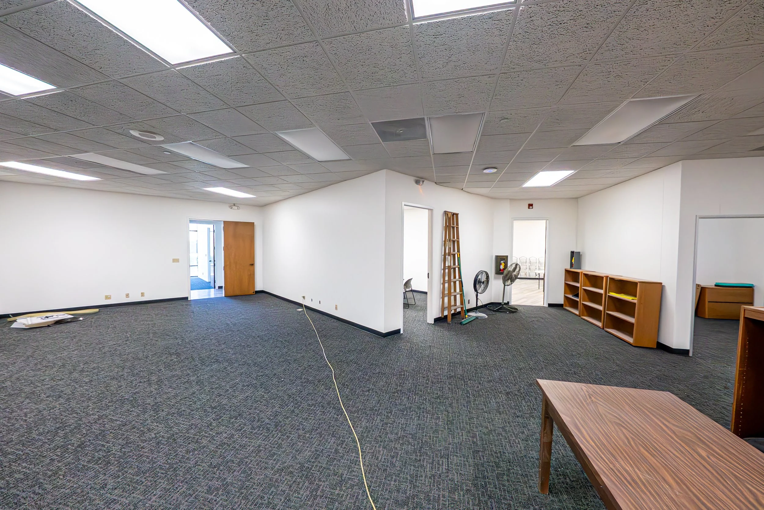 An empty office or classroom room with a dark gray carpet, white walls, and a ceiling with fluorescent lights. There are chairs, a wooden ladder, fans, and a small wooden shelf.