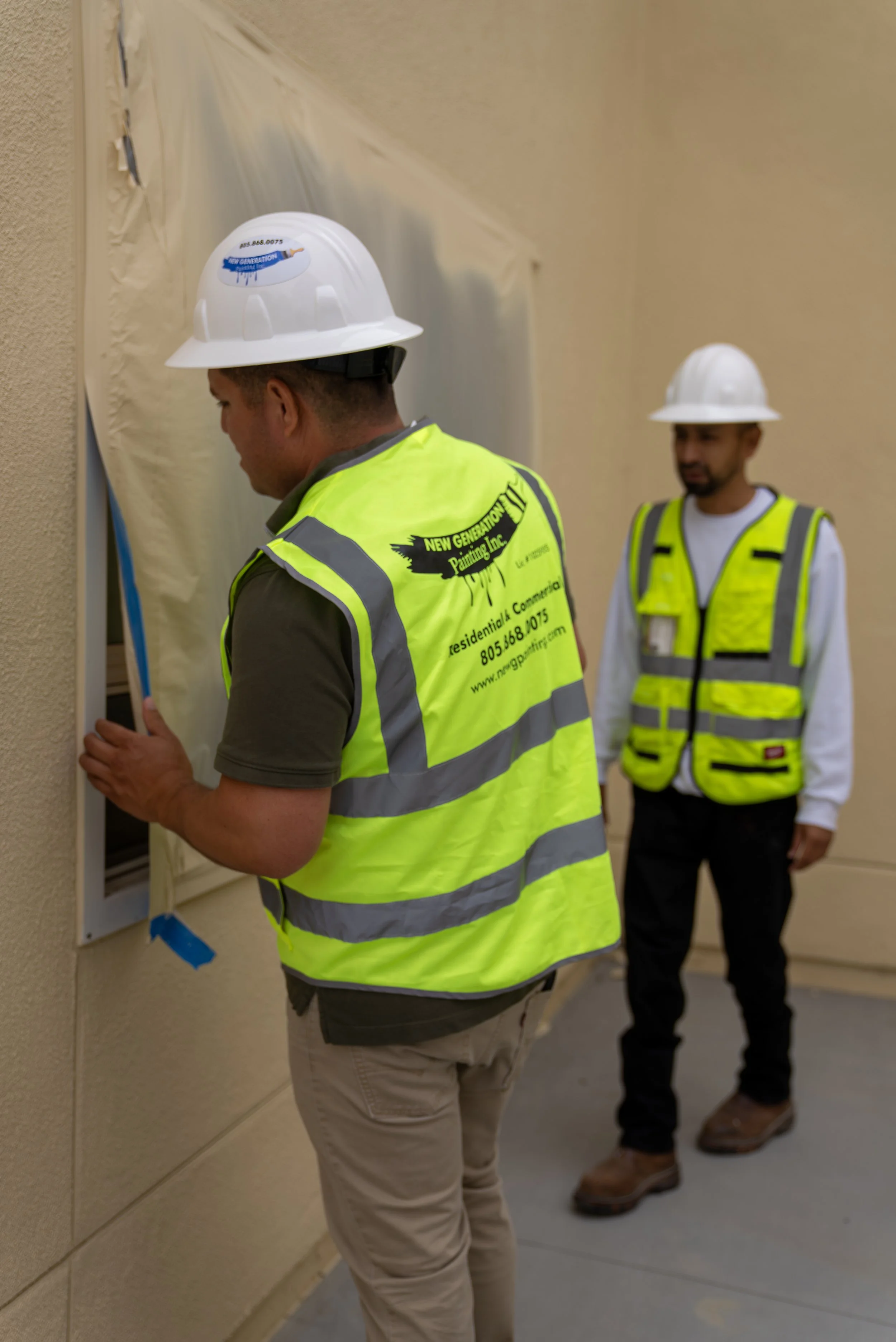 Two construction workers wearing white safety helmets and yellow safety vests working on a wall; one is closely inspecting a section while the other stands nearby, observing.