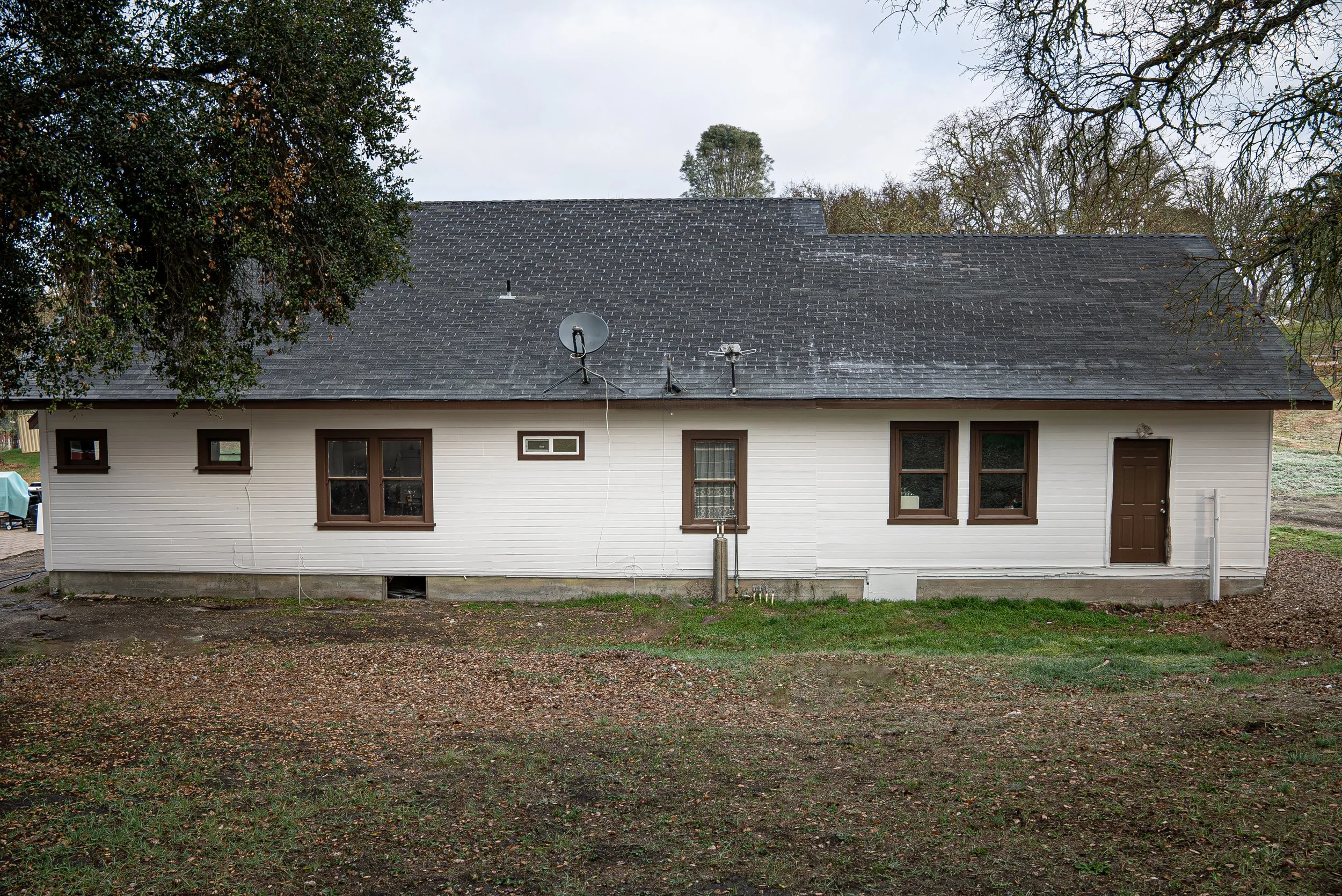 A house with a dark gray shingled roof, white siding, brown window frames, and a brown door, with trees and overcast sky in the background.