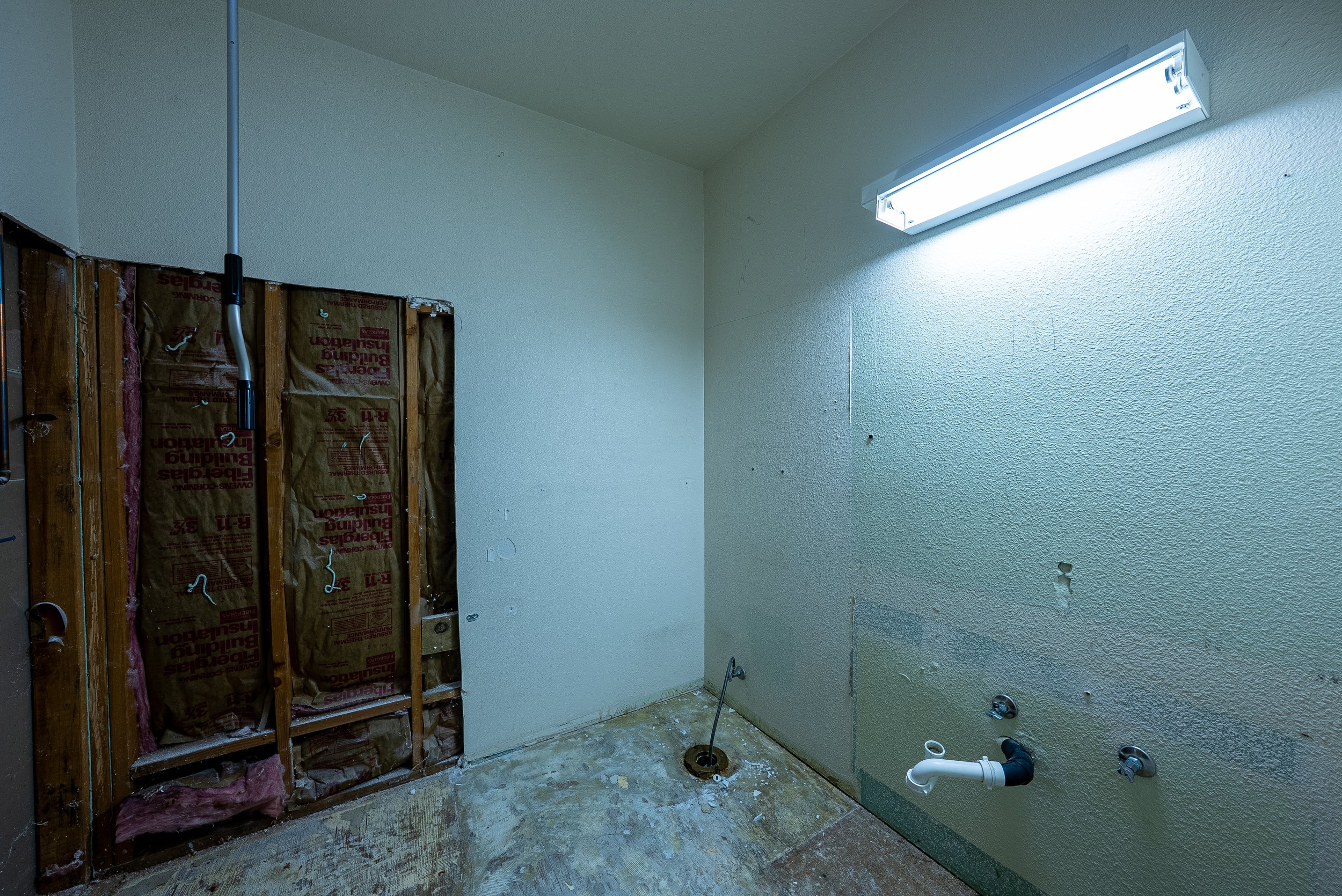 Partially renovated laundry room with exposed wall framing, insulation, and plumbing fixtures. Fluorescent ceiling light illuminates the space.