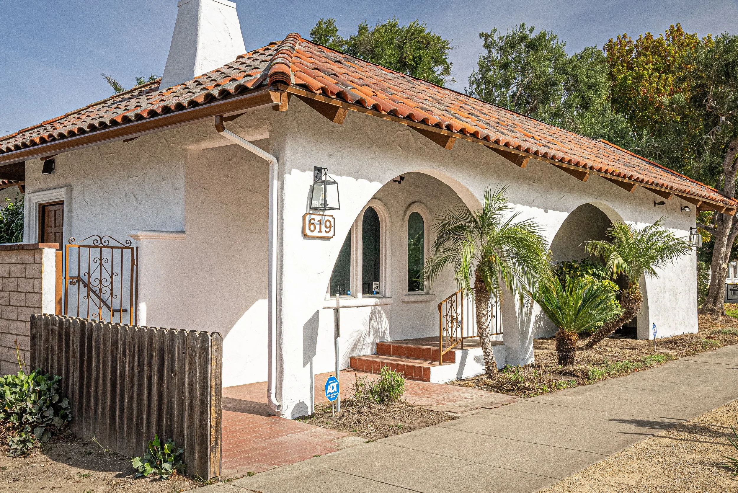 Front view of a white stucco house with a red tile roof, arched entryway, and decorative railing. There are palm trees and greenery in the yard, a wooden fence on the left, and an ADT security sign near the sidewalk.