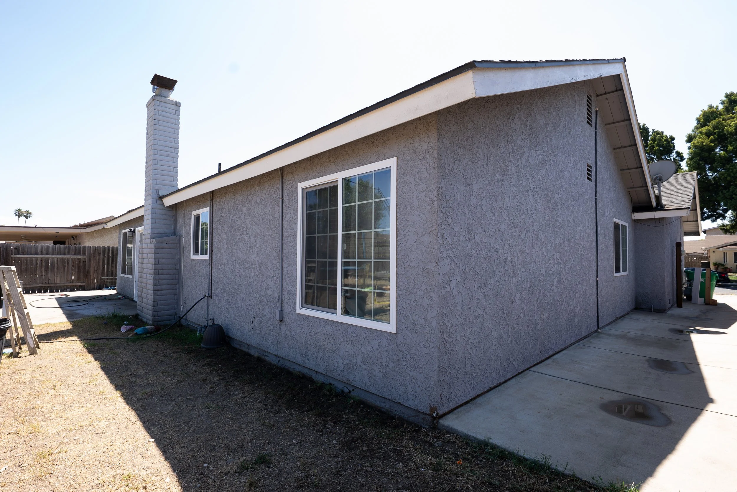 Backyard of a house with gray stucco walls, two large windows, a chimney, a concrete patio, and a wooden fence.