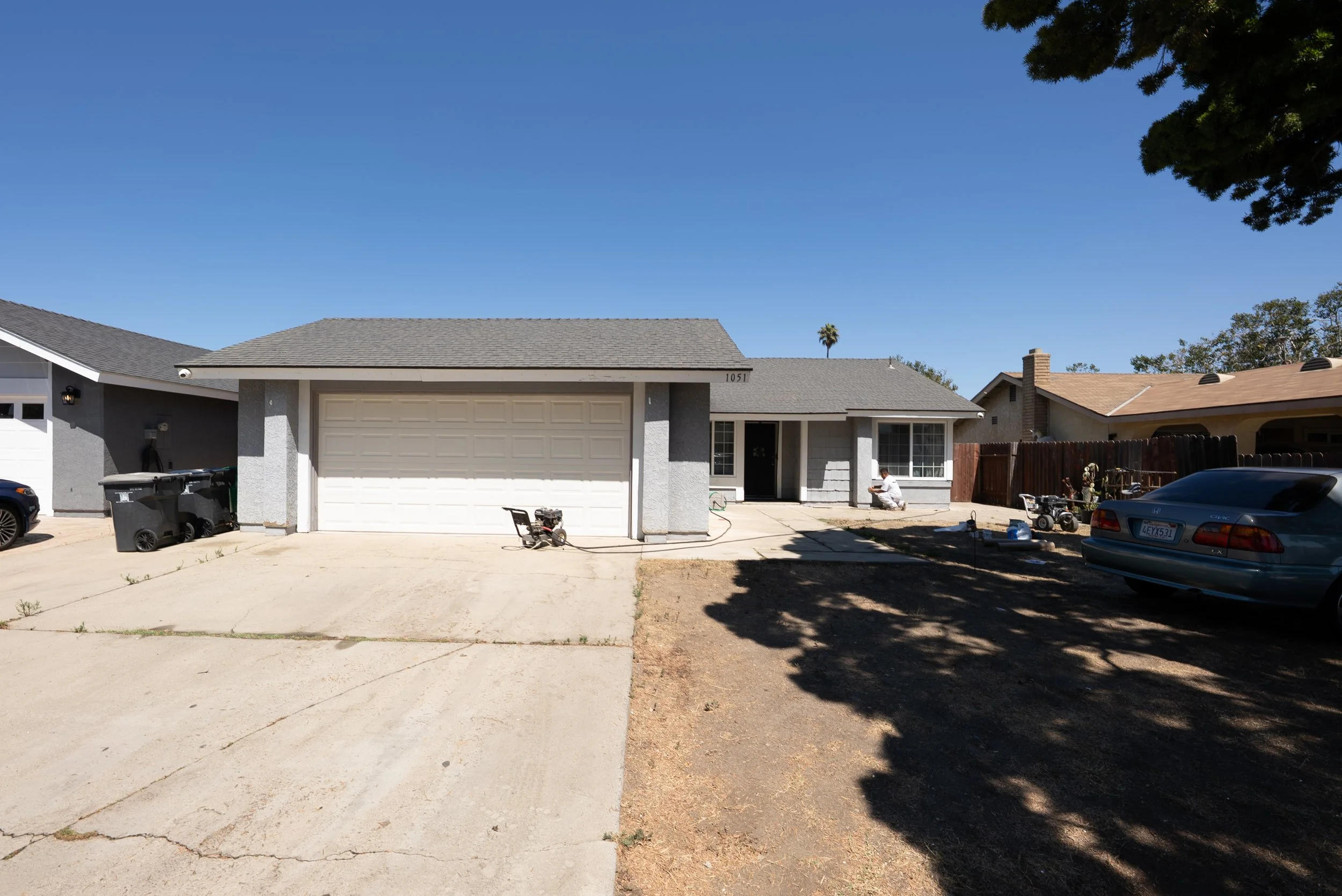 A single story gray house with a white garage door and a gray shingle roof in a residential neighborhood. There is a person kneeling near the house's window and several vehicles parked in the driveway. The yard has patches of dirt and grass, and a la