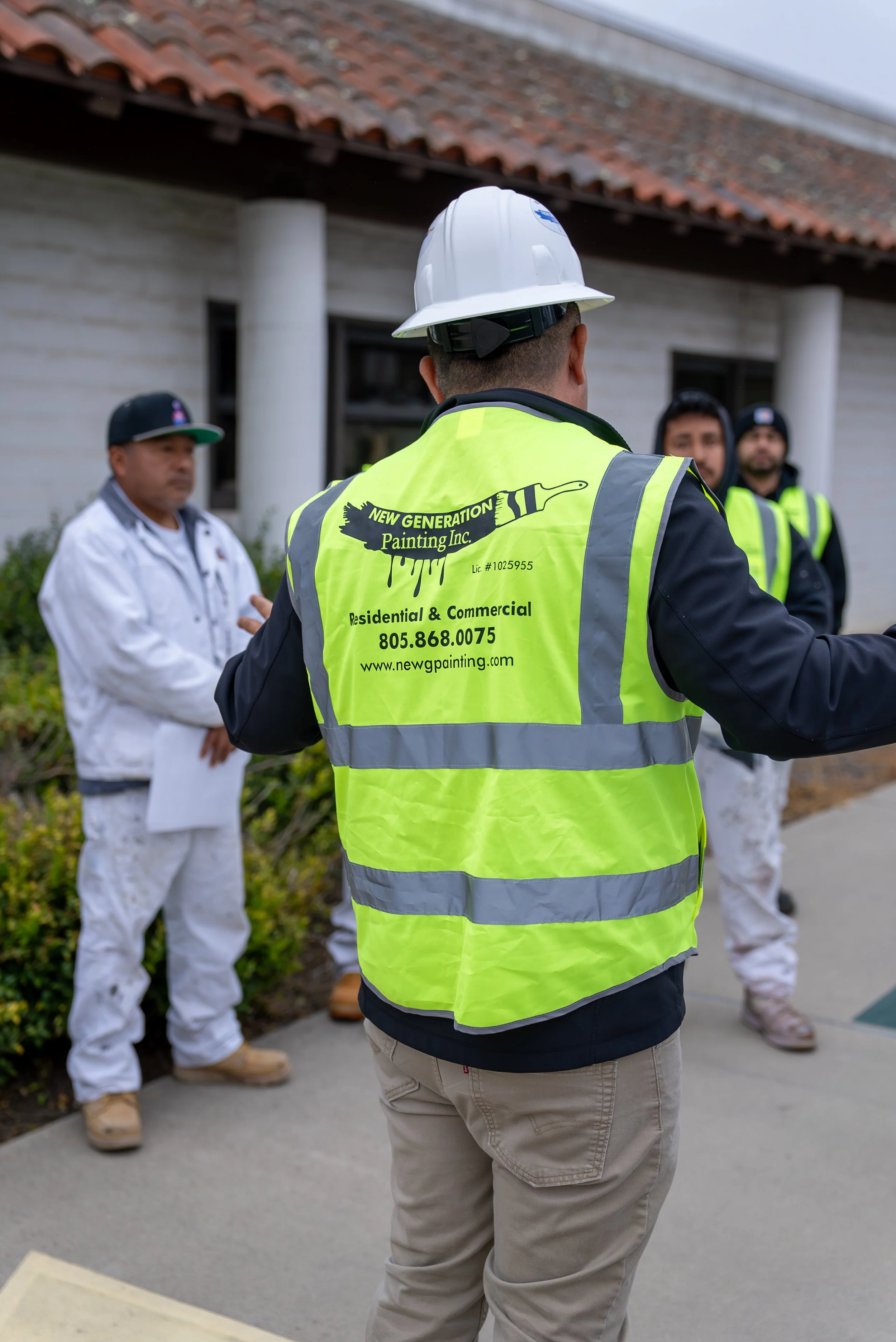 A construction worker wearing a bright yellow safety vest with the company logo and contact information, a white hard hat, and beige pants, standing outdoors in front of a house with group of men in the background.