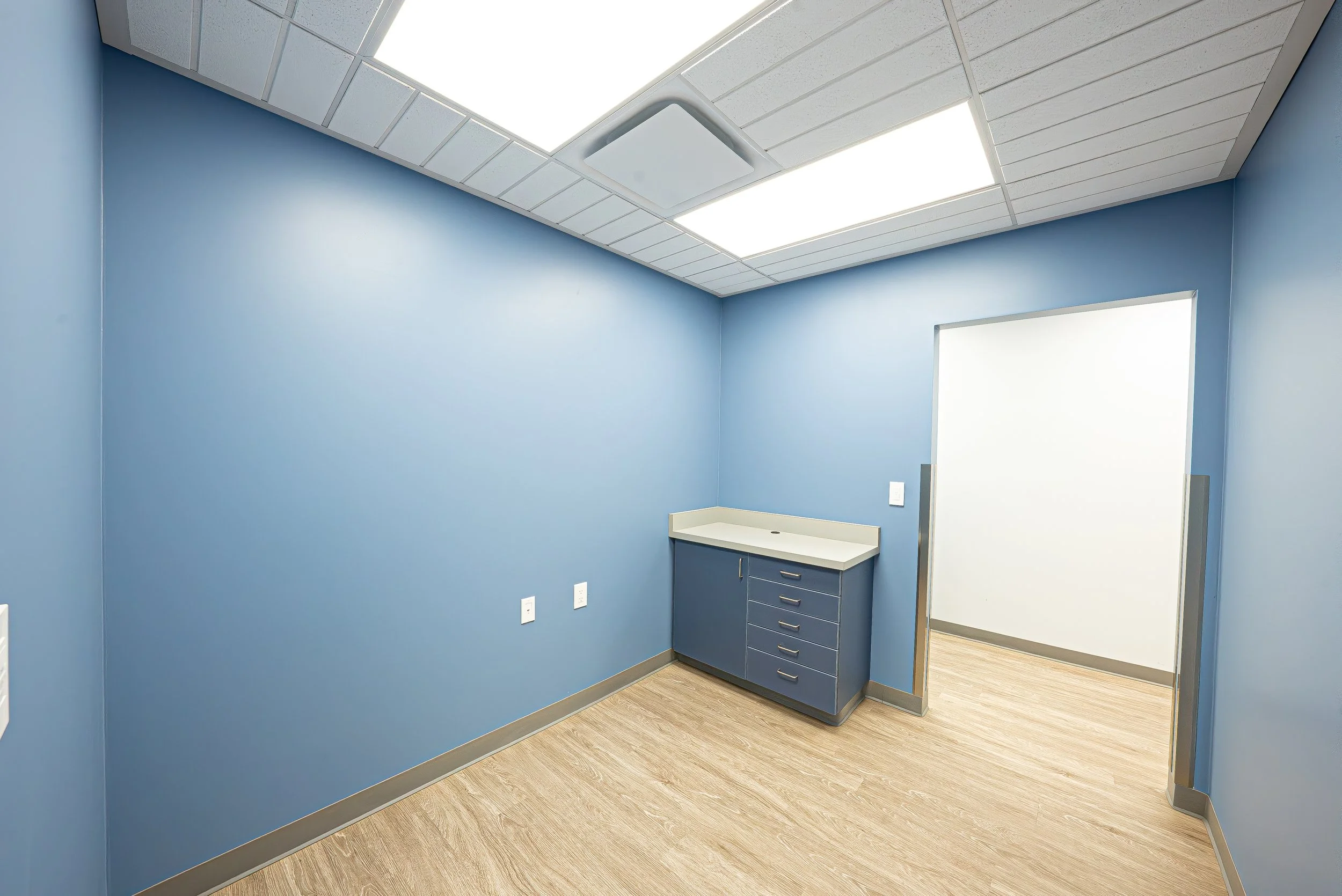 Empty medical examination room with blue walls, wood flooring, ceiling lights, and a cabinet near the door.