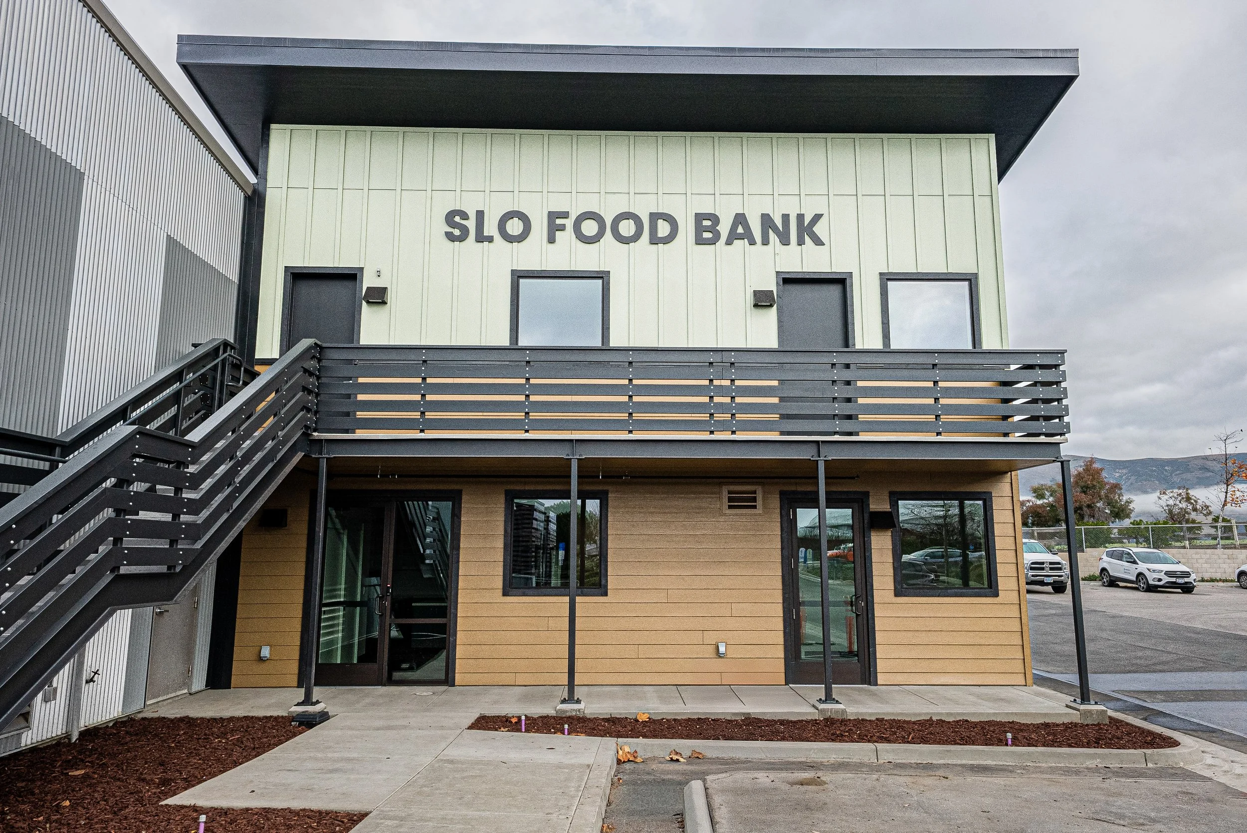 Exterior view of a modern building with a sign that reads 'SLO FOOD BANK'. The building has a combination of metal siding and wood paneling, with black-framed windows and an external staircase. The sky is cloudy and there are a few parked cars in the