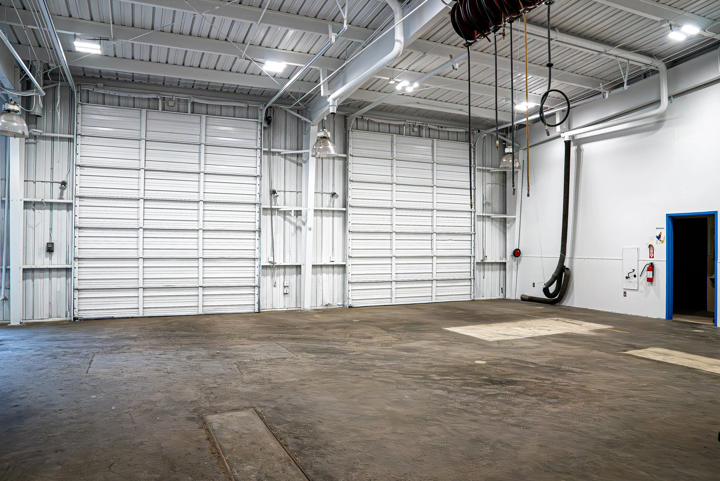Empty industrial warehouse or garage with metal walls, ceiling, and a concrete floor, along with industrial lighting and ventilation systems.