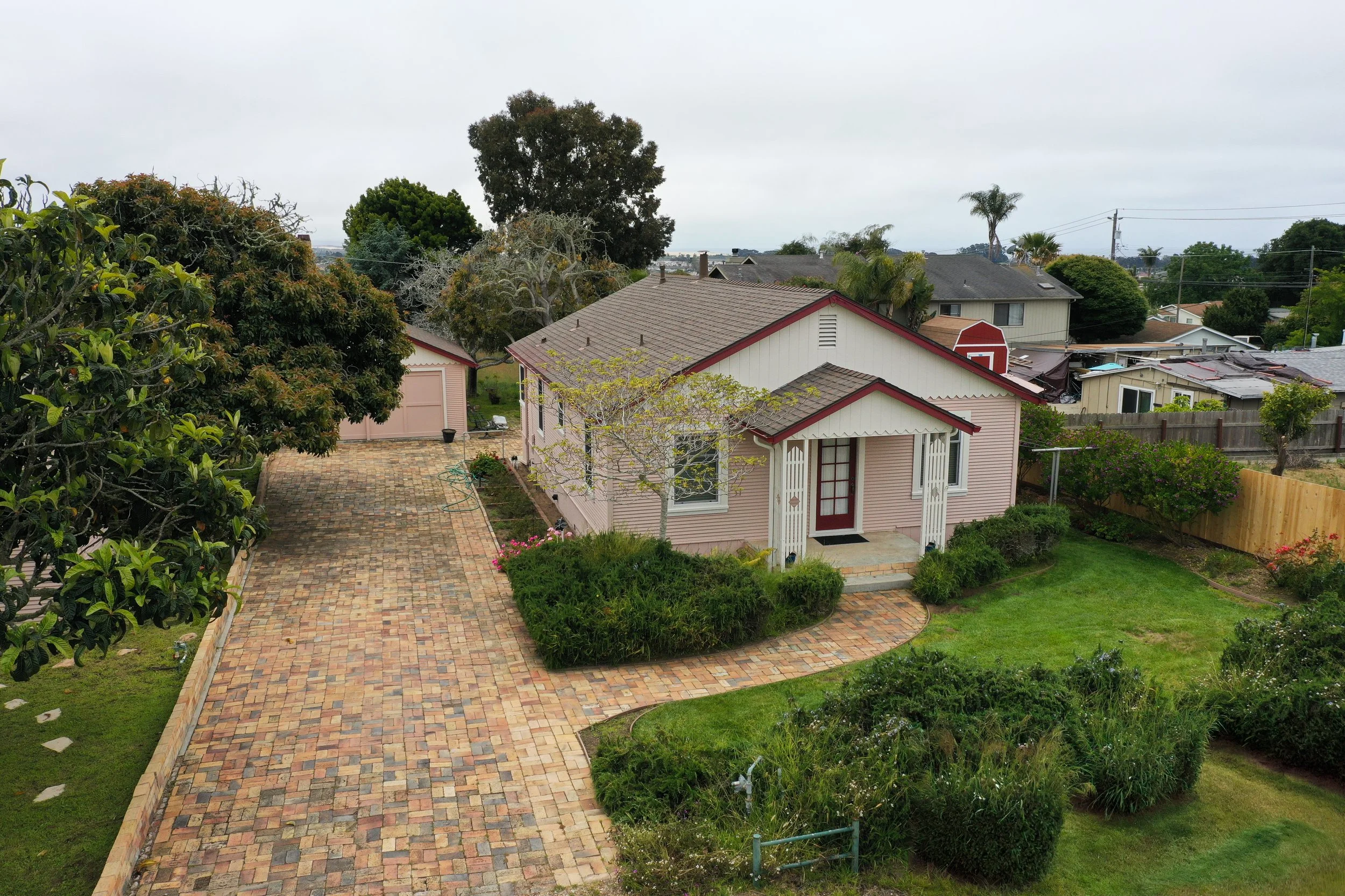 A backyard with a paved brick pathway, a small pink house with a red door, green shrubs, trees, and neighboring houses under an overcast sky.