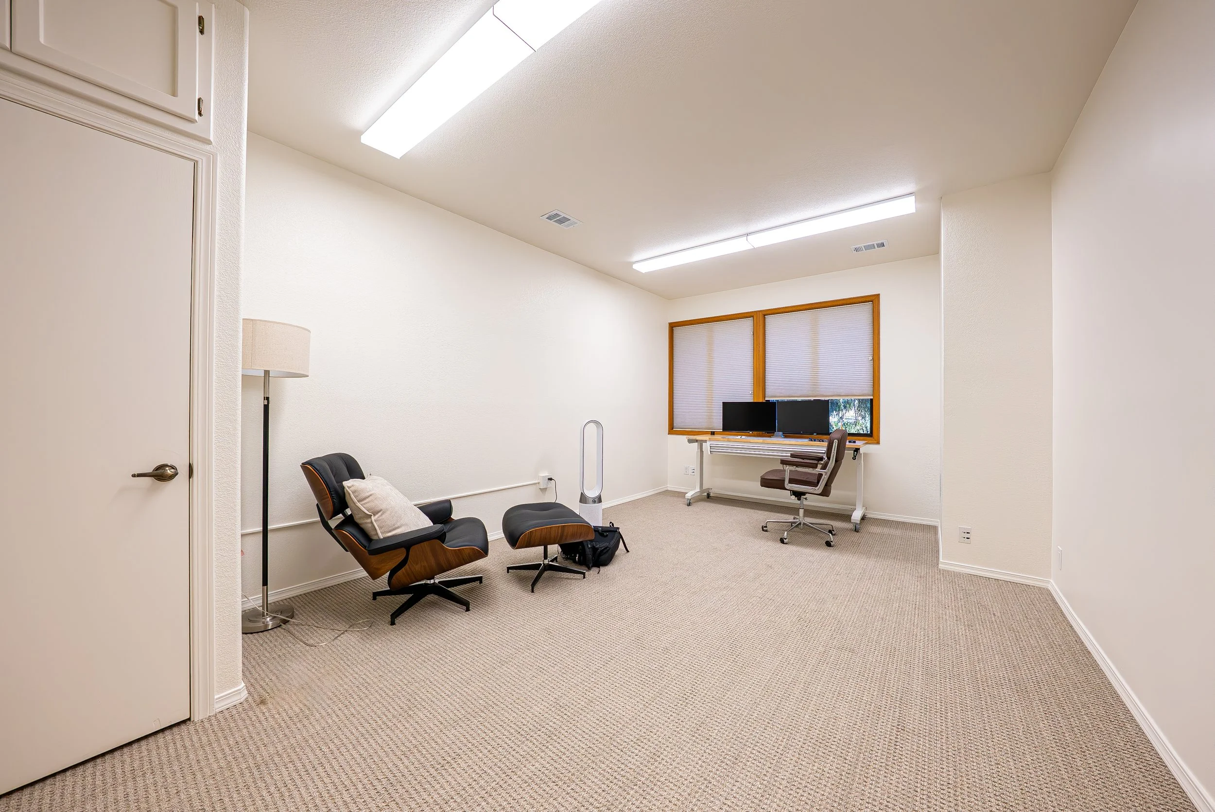 A small office with beige carpet, white walls, and two fluorescent ceiling lights. There is a wooden window with closed blinds. In the room, there are two black chairs with wooden armrests, one with a pillow, a desk with two computer monitors, a brow