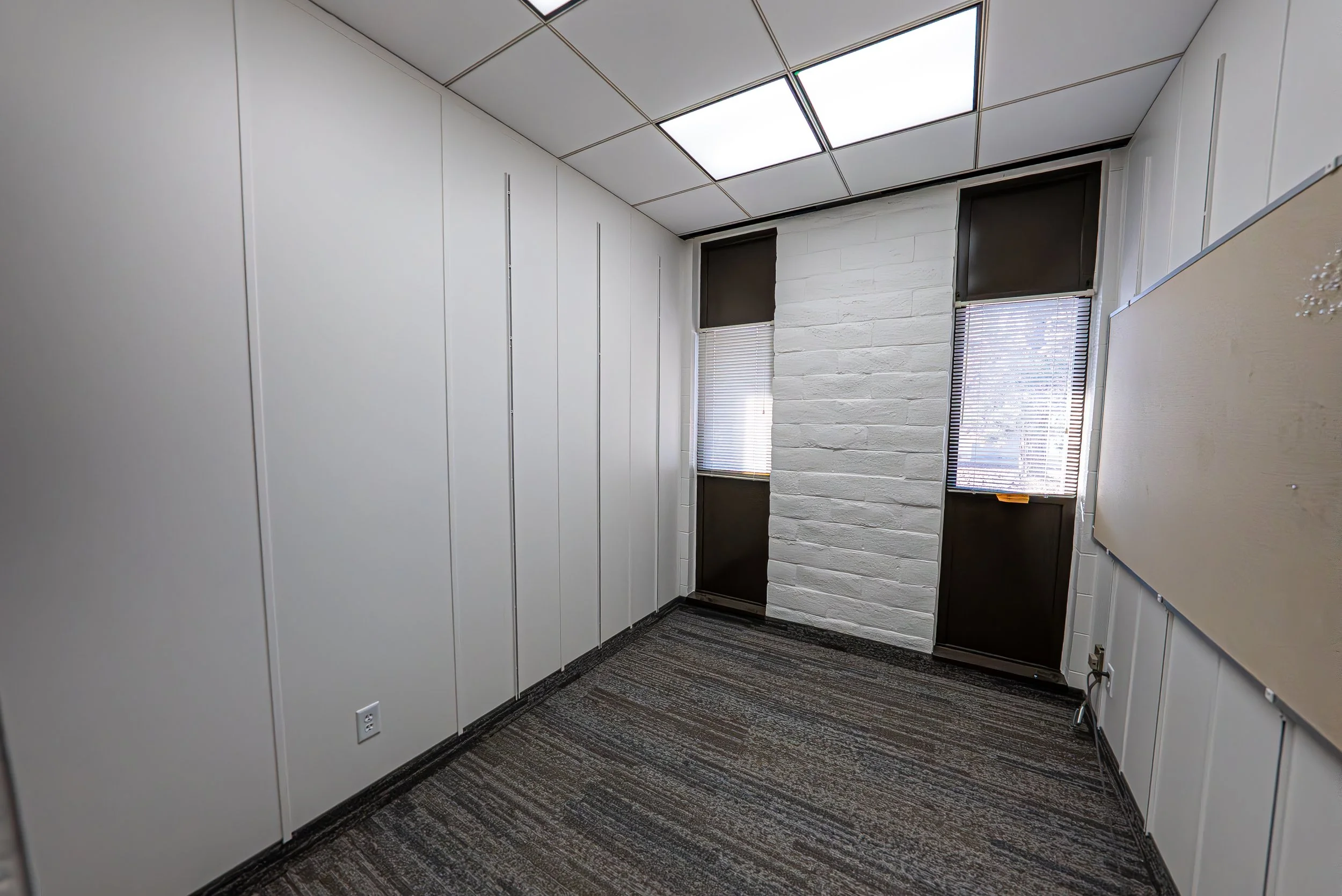Empty office corner with white walls, black window blinds, white brick accent wall, and carpeted floor.