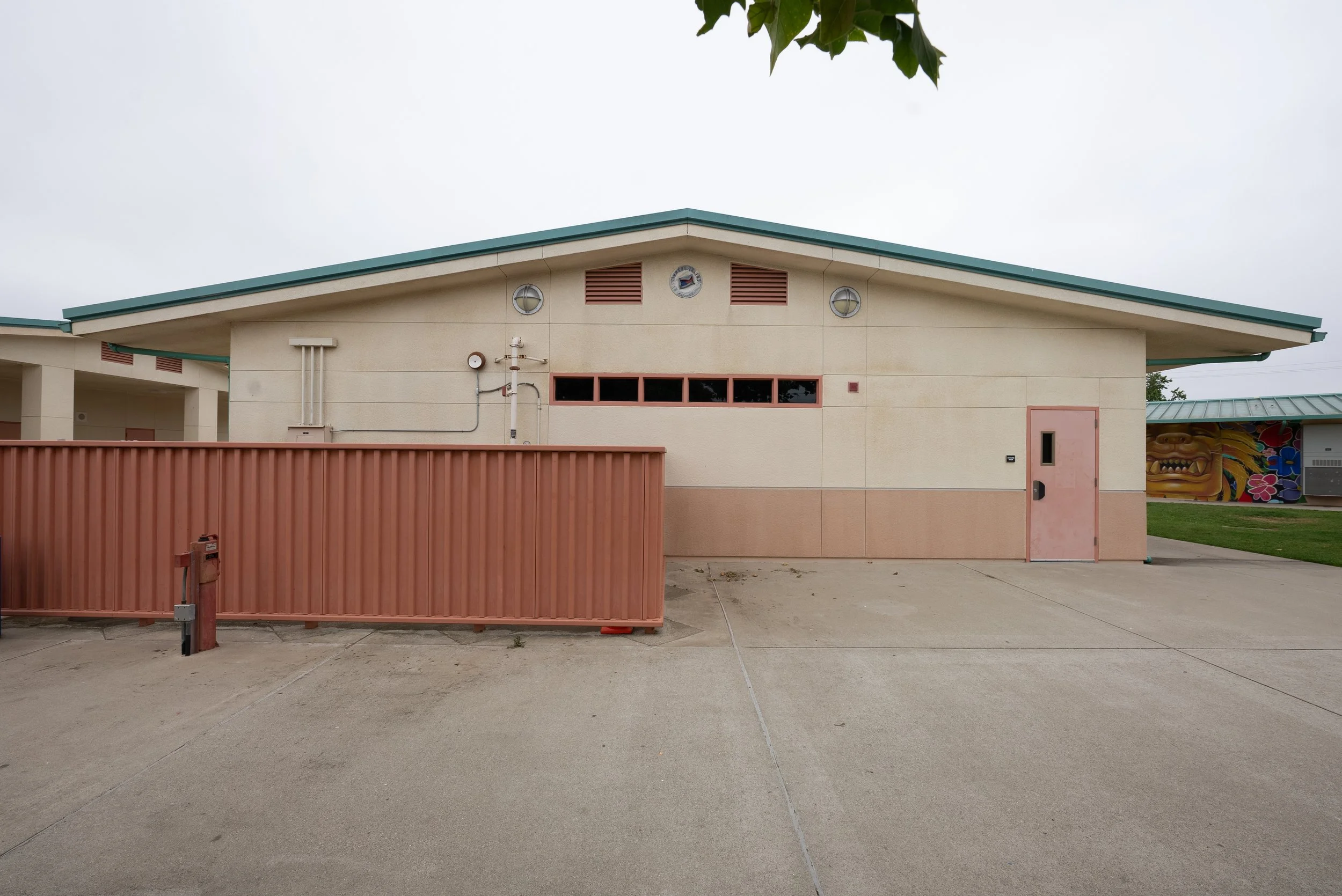 A beige building with a green roof, small windows, and a wall-mounted clock. A red fence and a sidewalk are in the foreground, with overcast sky in the background.
