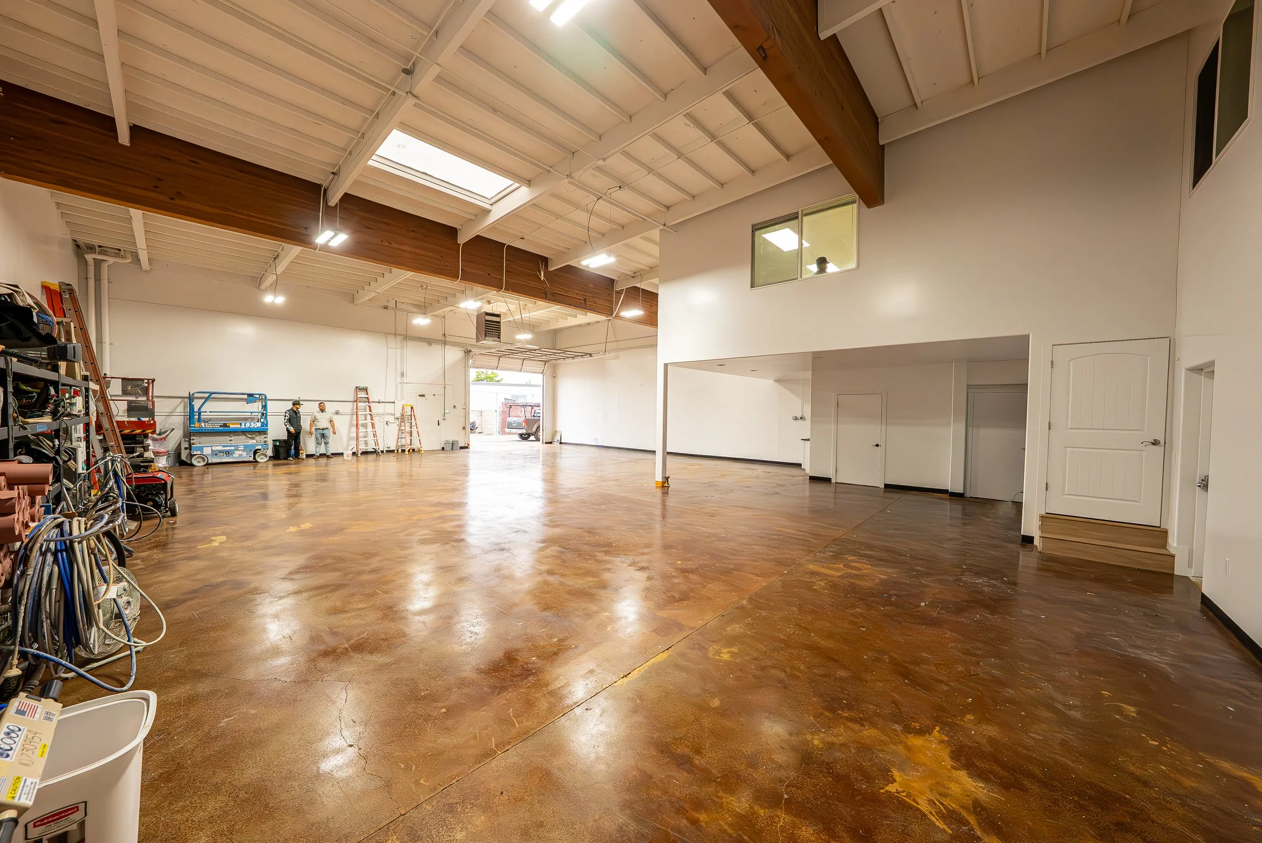 Interior of a spacious warehouse or workshop with a high ceiling, skylights, and wooden flooring. Two people are standing near the back, some ladders and equipment are visible on the left side, and a vehicle can be seen outside through the open back 