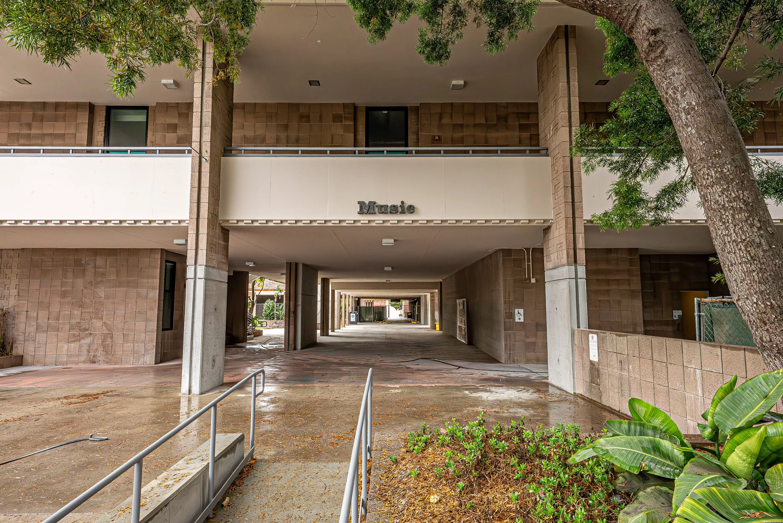 Multilevel parking garage with a sign that says 'Music' on the upper level. There are trees, plants, and a walkway in front of the garage.