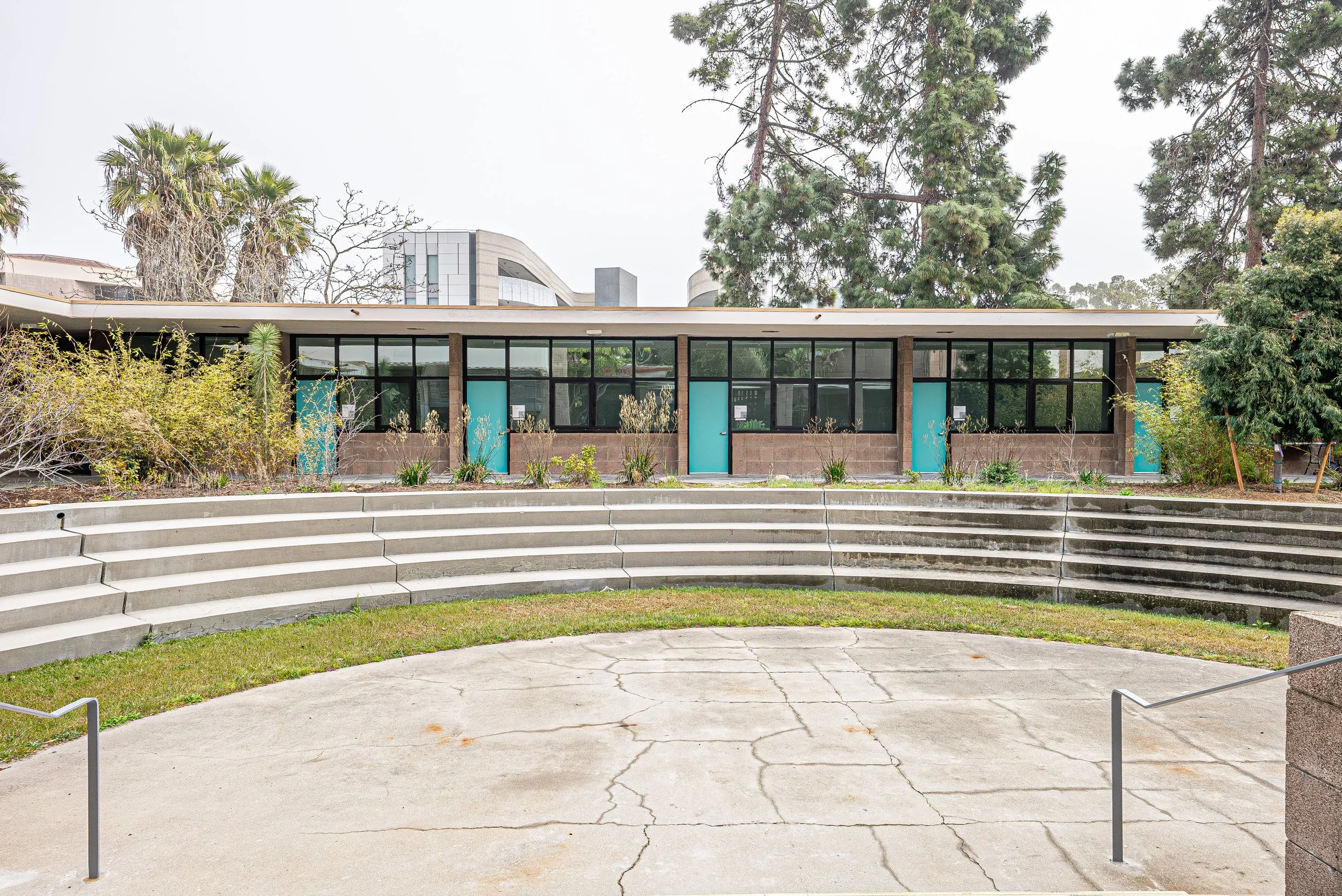Outdoor amphitheater with curved concrete steps leading up to a stage area, surrounded by bushes, trees, and a modern building in the background.