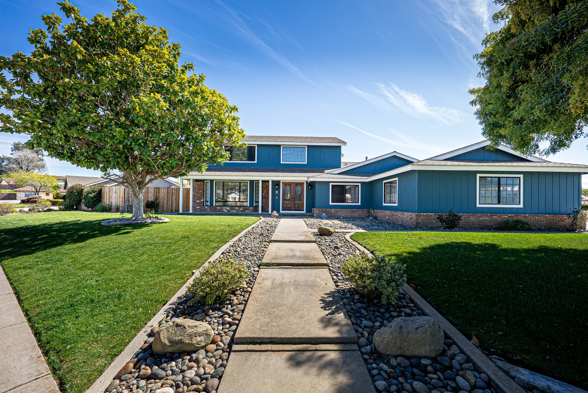 A blue two-story house with a front sidewalk, surrounded by a manicured lawn and large trees, under a clear blue sky.