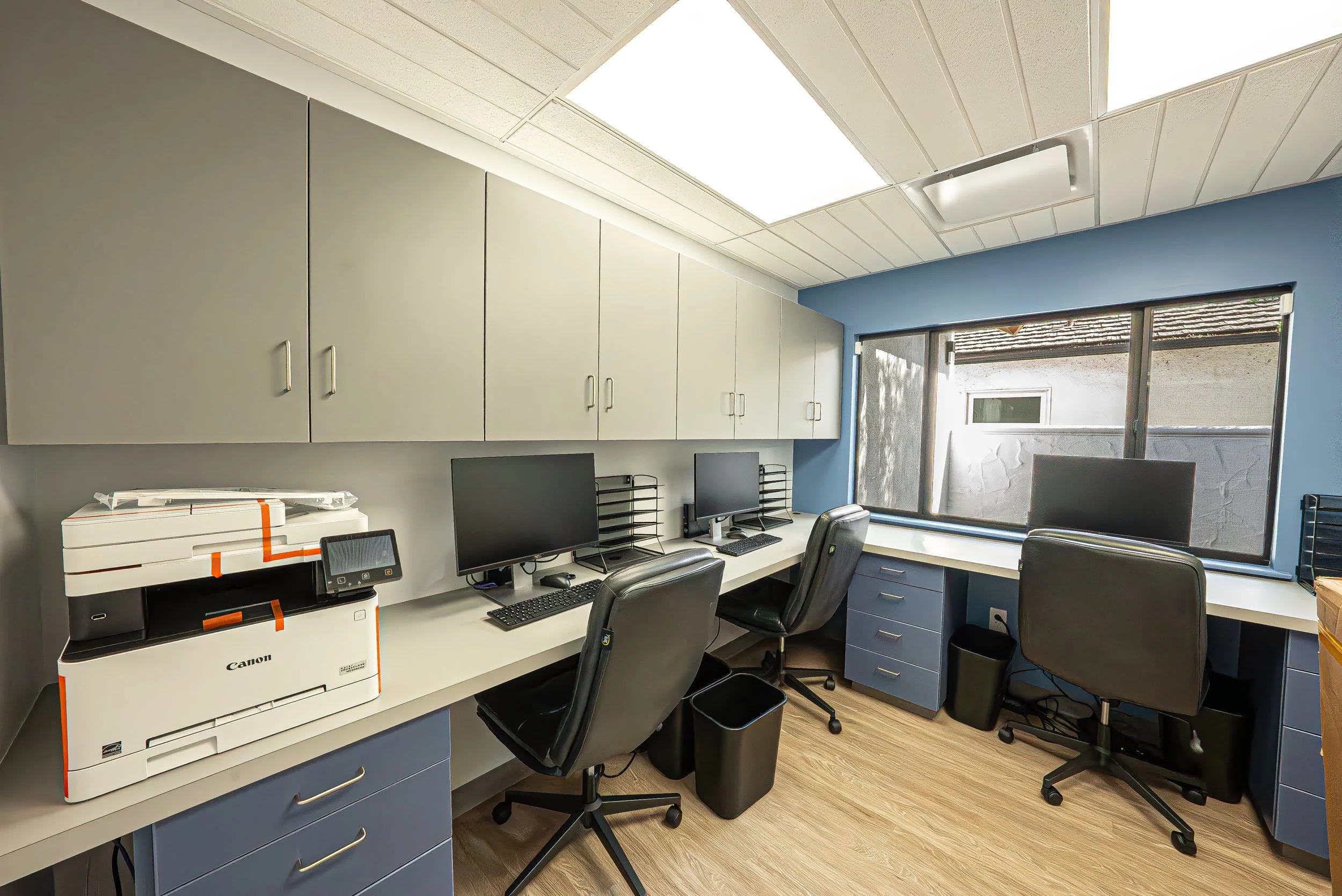 An office workspace featuring a white desk with a Canon copier, two computer monitors, and office chairs. Cabinets are mounted on the wall above the desk. A window lets in natural light, revealing rooftops outside.
