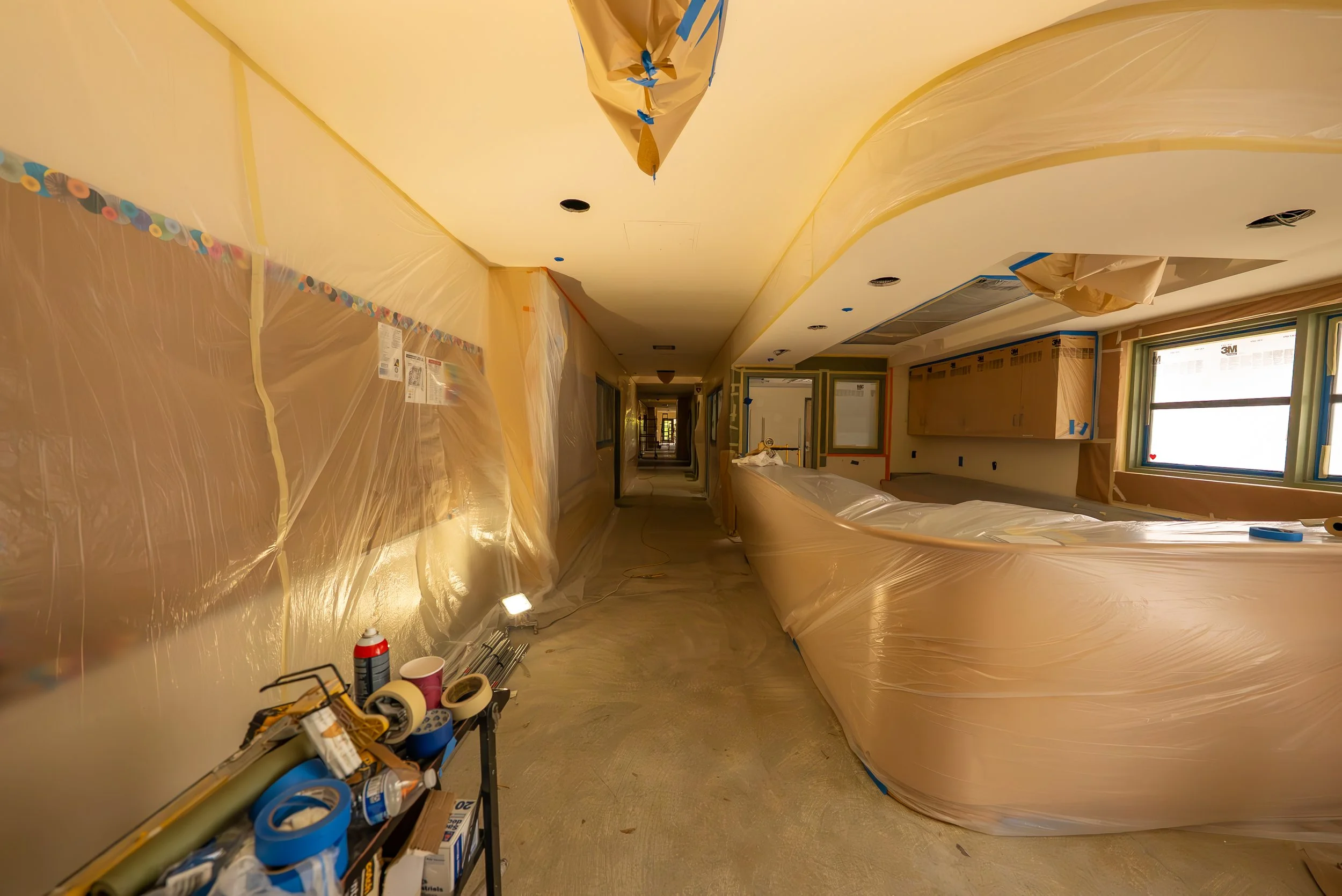 Hallway under construction with plastic sheeting covering the walls and ceiling, construction tools and supplies on the floor and shelves, and a window allowing natural light in.
