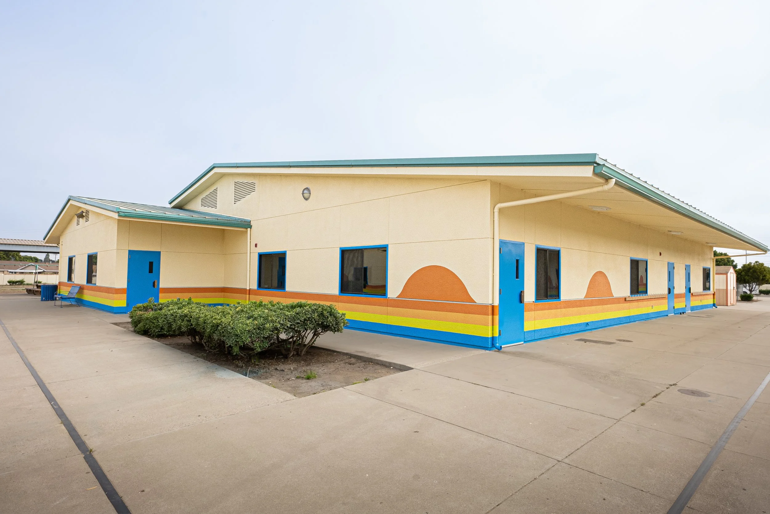 A colorful single-story building with a rainbow painted on the lower part of the walls, blue doors and windows, a light yellow exterior, and a green roof, situated in a paved area with some bushes and a bench in front.