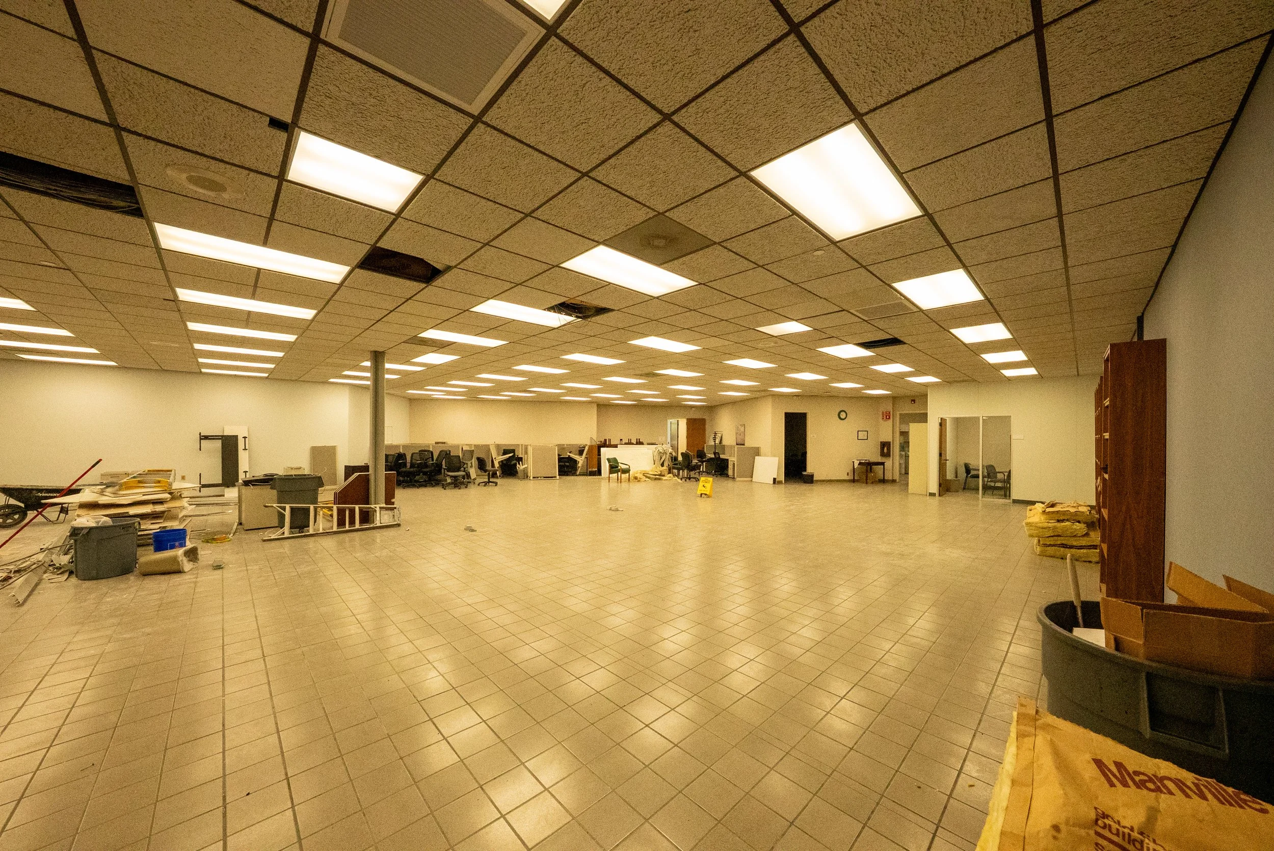 A large, empty room with a tiled floor and a drop ceiling with fluorescent lights. Some ceiling tiles are missing or damaged. There are scattered items and debris around, including trash bins, boxes, and furniture, indicating ongoing renovation or cl