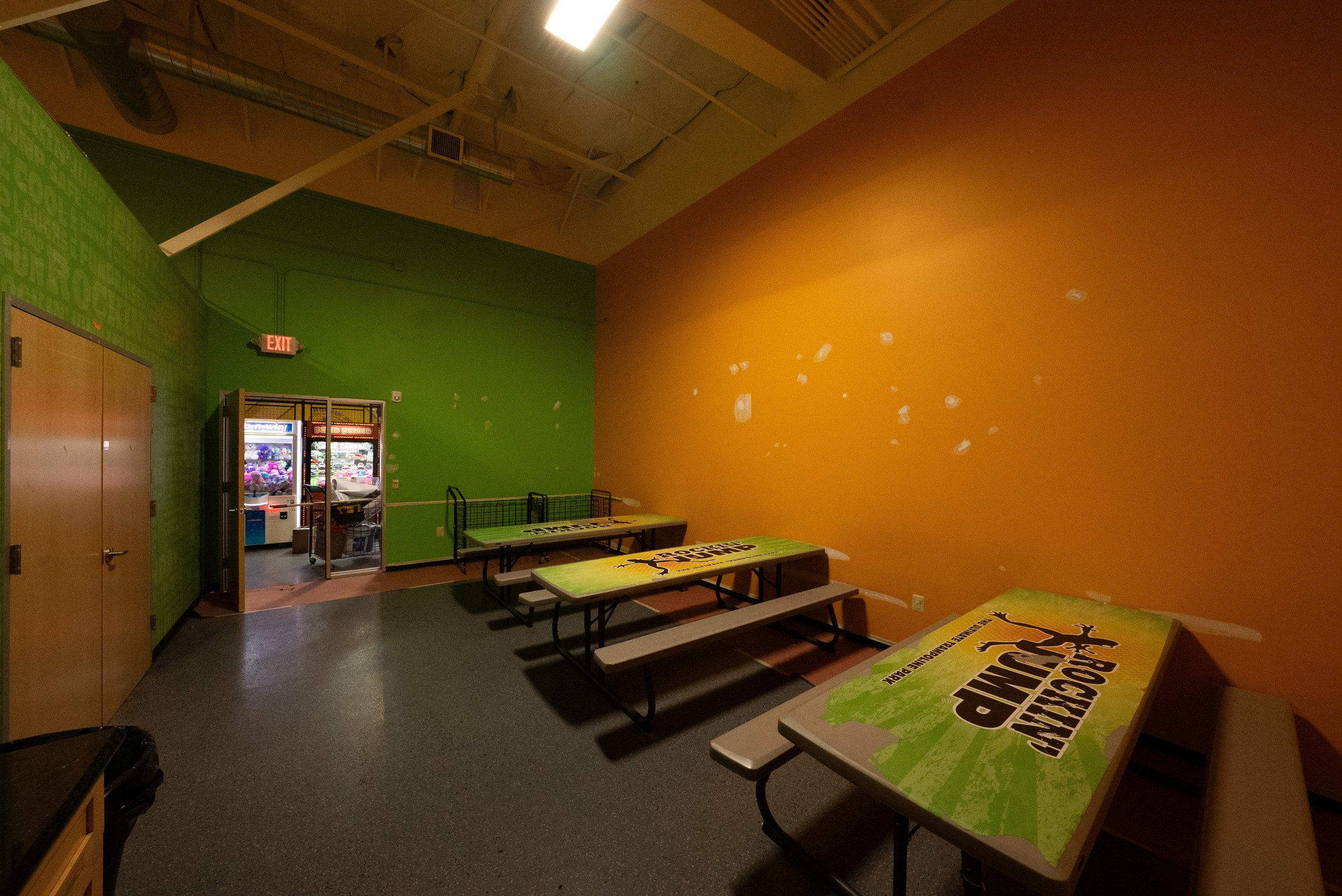 Indoor arcade or game area with green and orange walls. Several picnic tables with a sports-themed print. A vending machine is visible near a door labeled 'EXIT'.