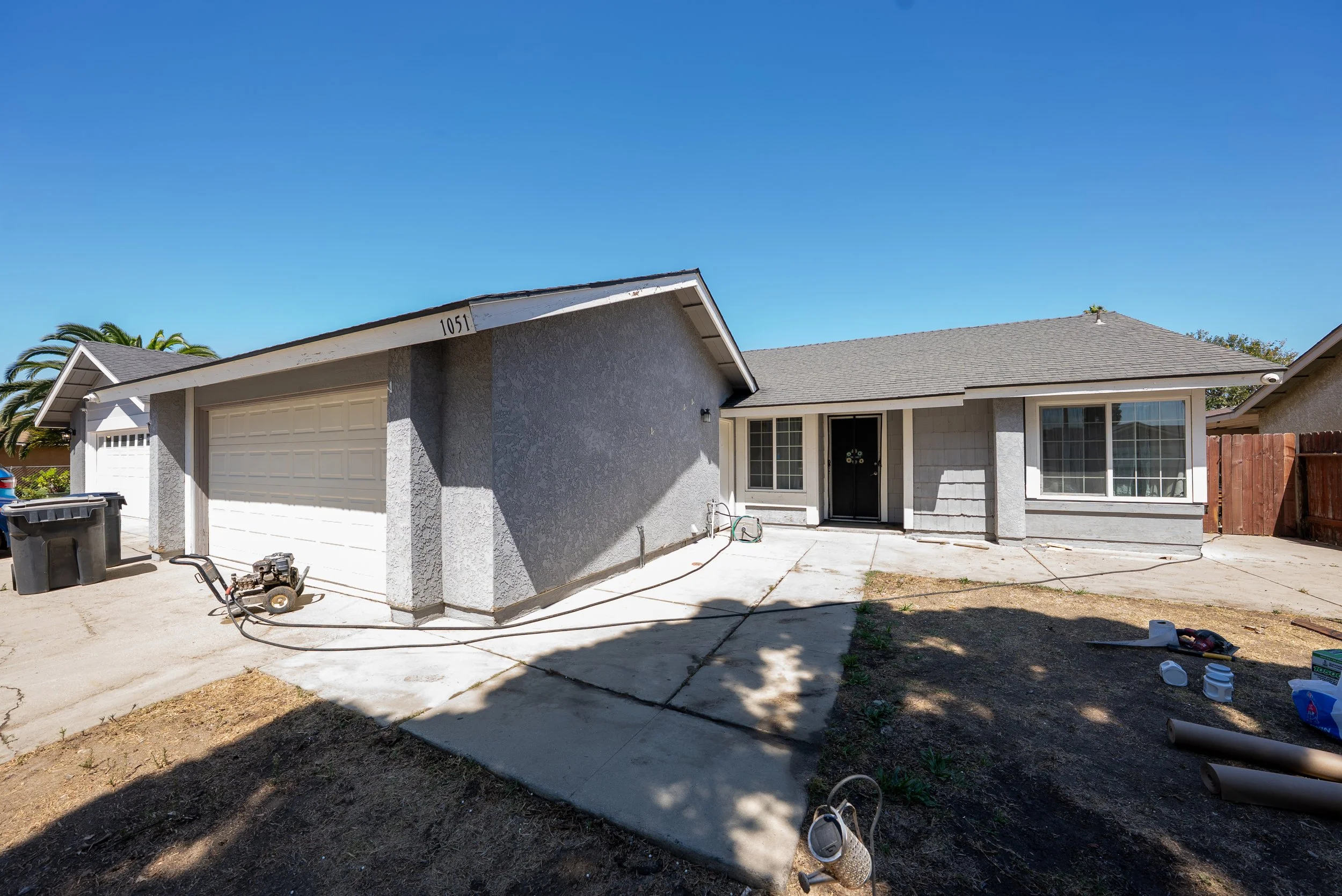 The image shows a suburban house with a gray exterior, white trim, and a gray shingle roof. There is a front door with a wreath and large front windows. The driveway has construction tools and supplies, including a pressure washer, a trash bin, and c