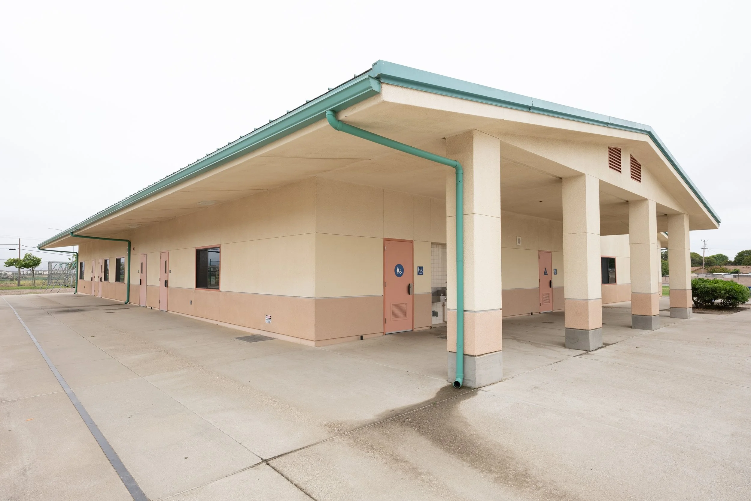 A beige building with a turquoise roof and mortar pipes, windows, and bathroom doors outside on a concrete parking lot.