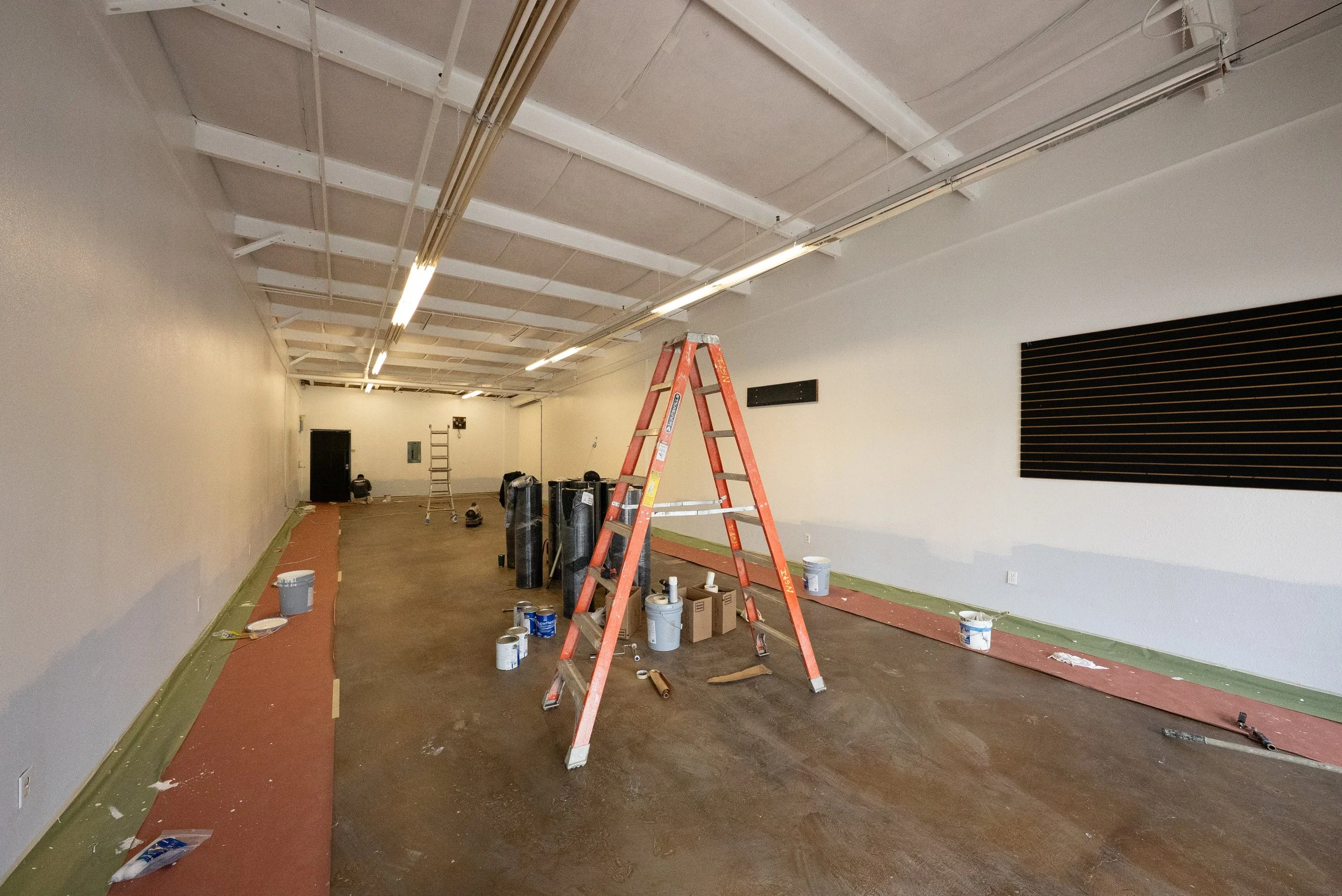 A large indoor space undergoing renovation with construction tools and supplies, including a red ladder, paint cans, and a workwoman in the background.