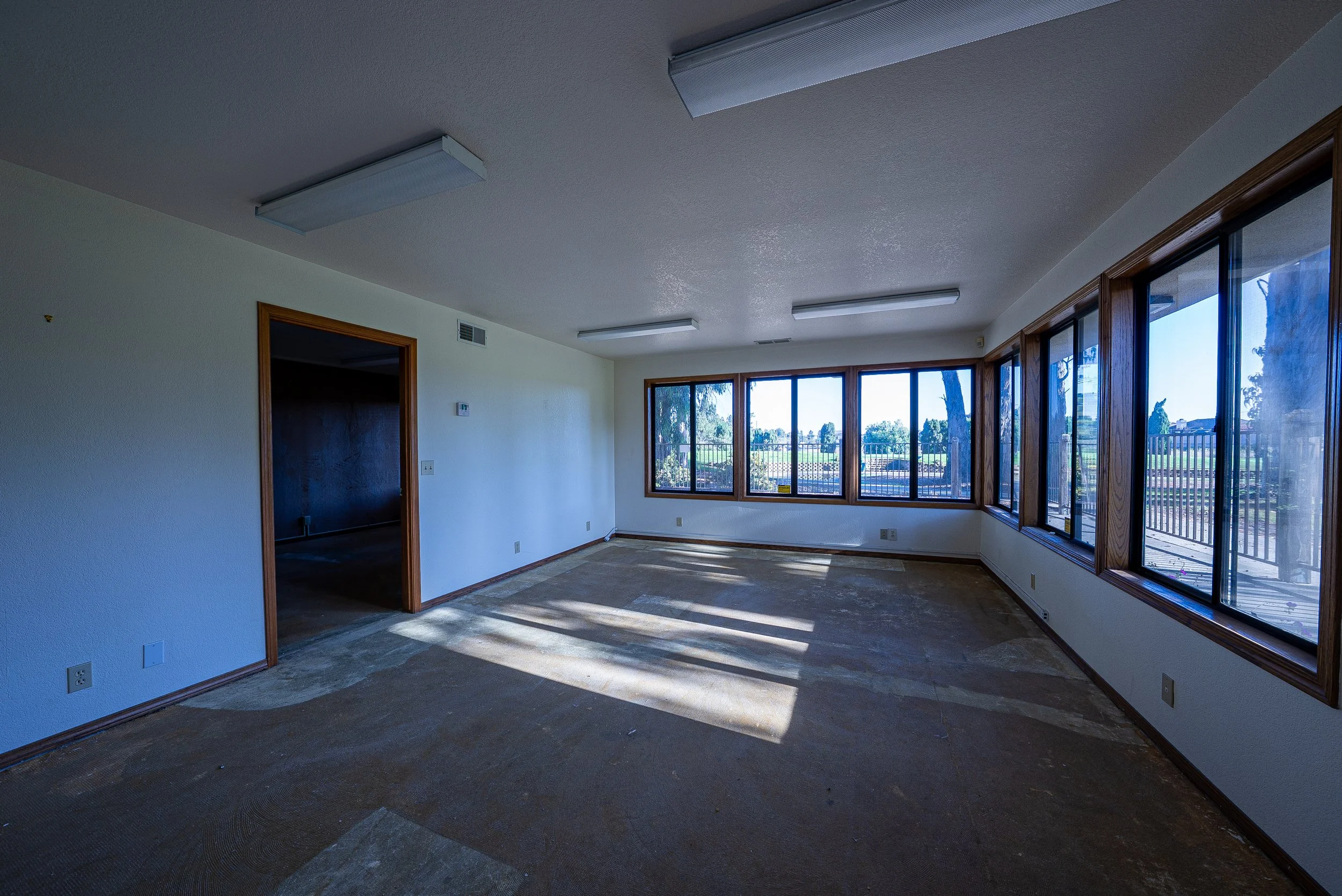 An empty room with large windows, a sliding door leading outside, and ceiling lights, letting in natural light.
