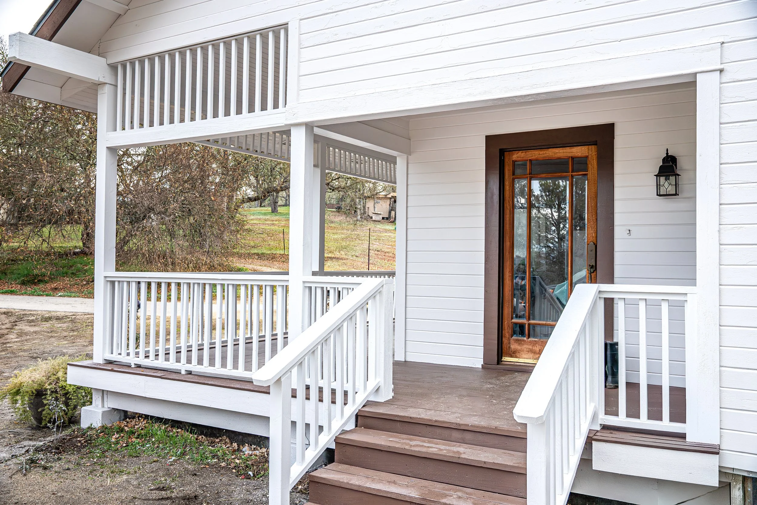 Front porch of a white house with brown steps and a wooden door, black lantern style light fixture, and a small porch railing with a leafless tree and yard in the background.