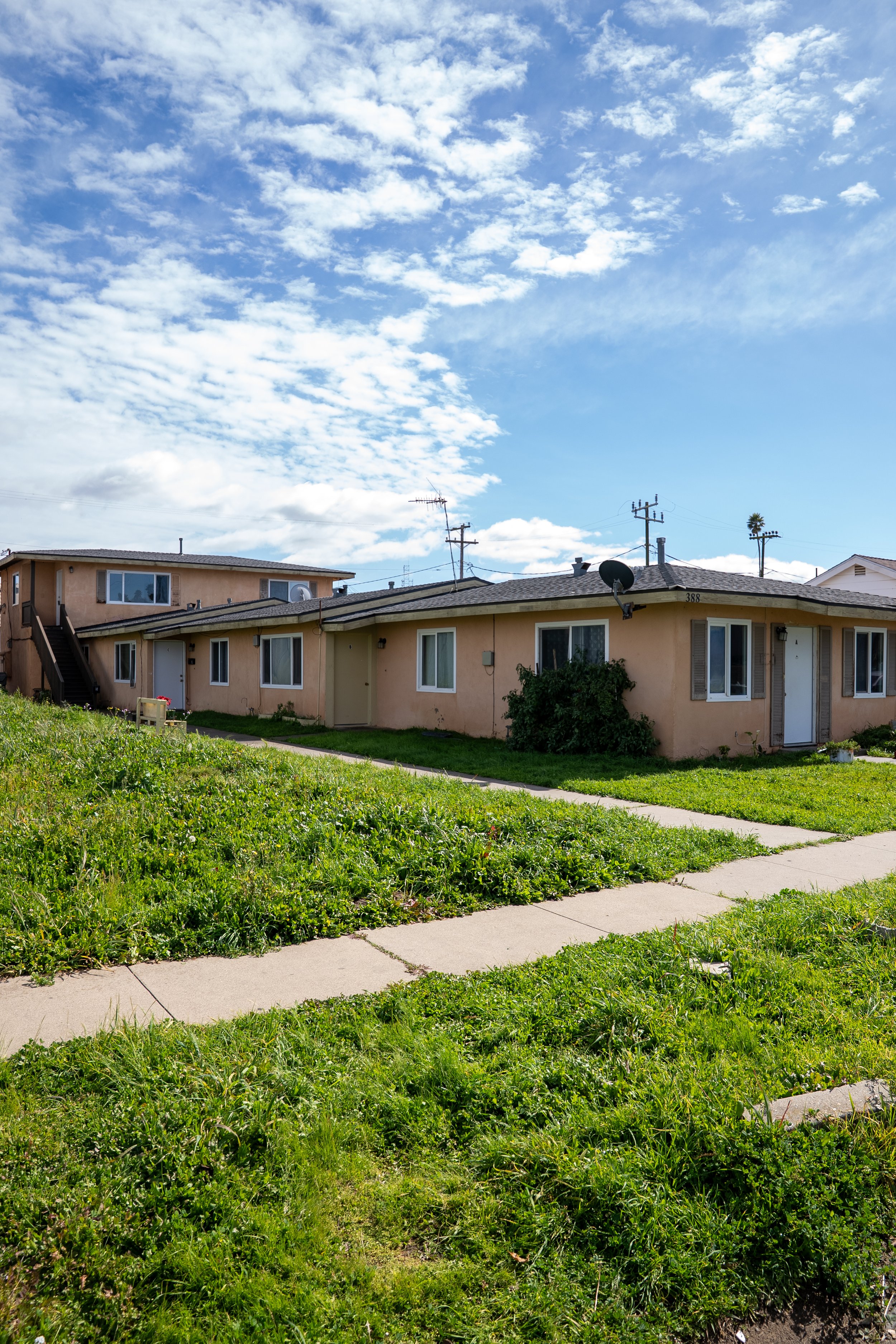 A row of beige apartment buildings with multiple windows and doors, surrounded by green grass, with a concrete sidewalk in front. The sky is mostly clear with scattered white clouds and utility poles with wires in the background.