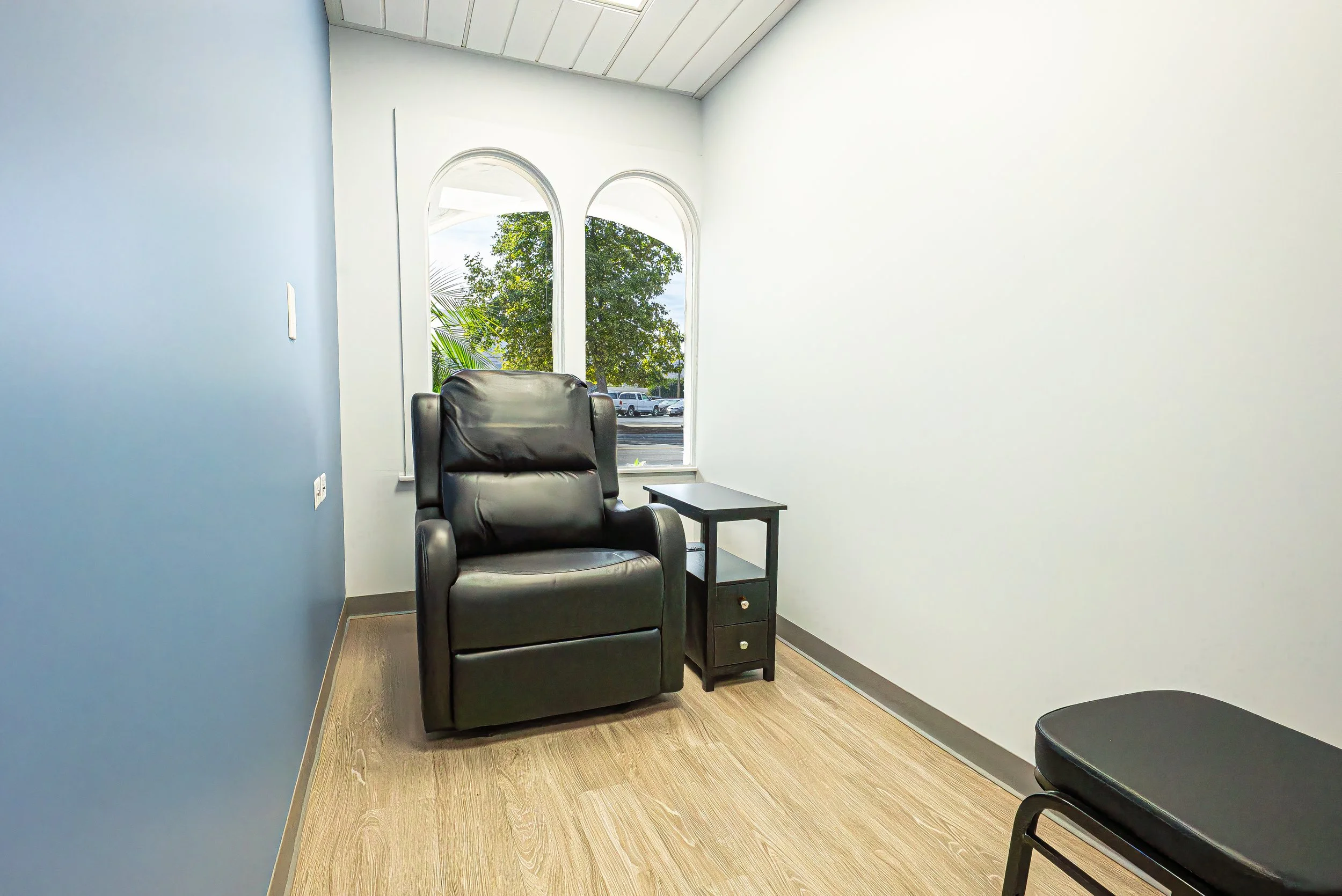 An office with a black leather armchair, a small black side table, and a window showing trees outside. The room has white and blue walls with a wood floor.