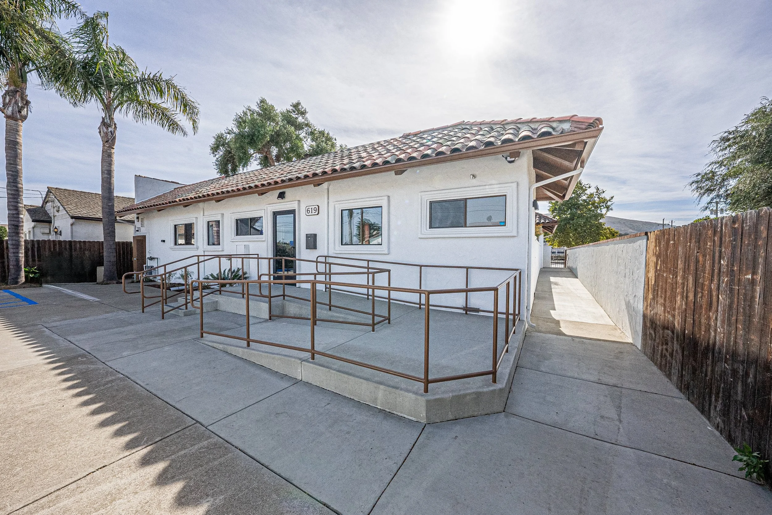 White building with a sloped tiled roof, windows, a ramp for accessibility, and a sidewalk, with tall palm trees and a wooden fence nearby under a partly cloudy sky.