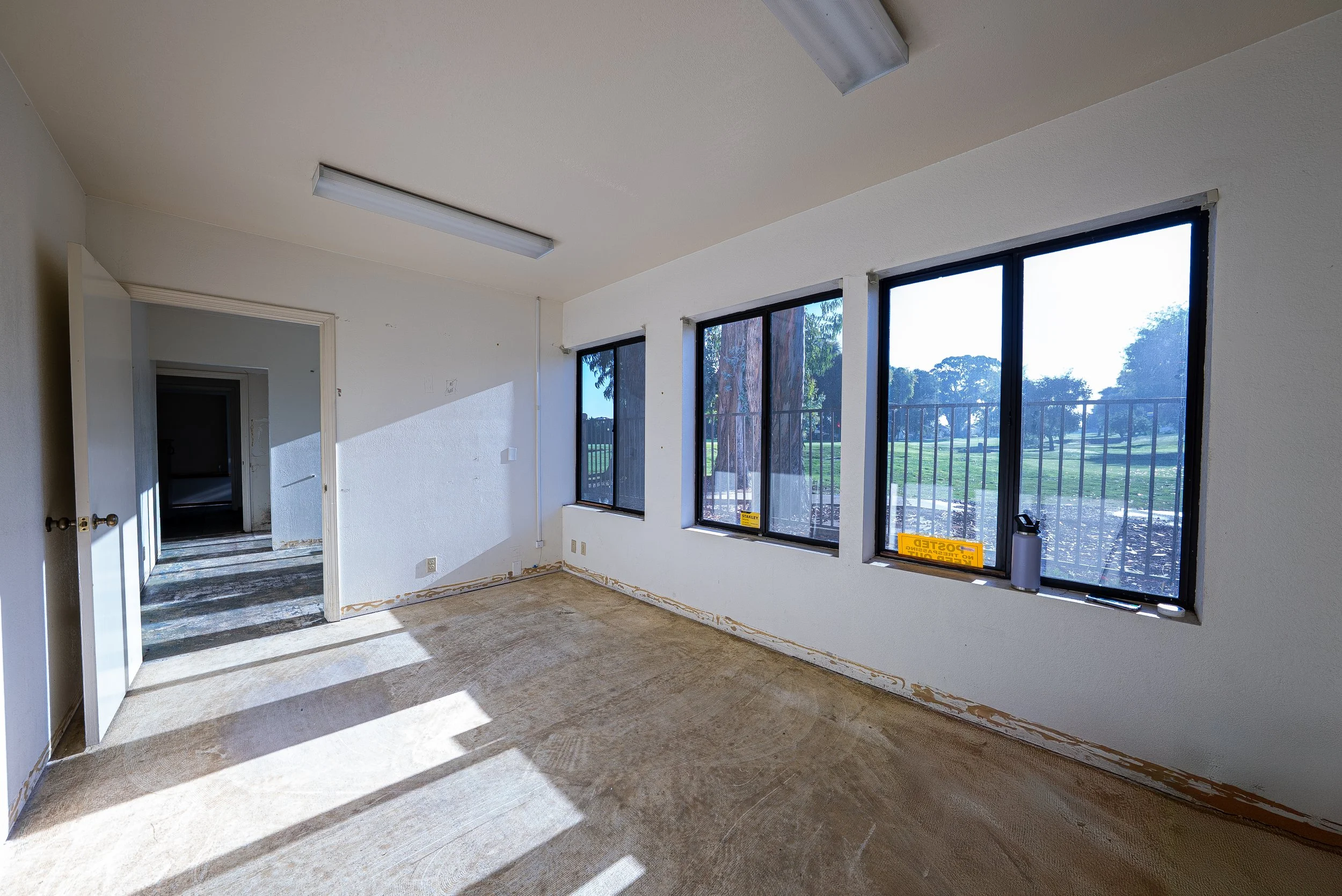 Empty room with large windows showing trees outside, sunlight casting shadows on the floor, and a door leading to another room.