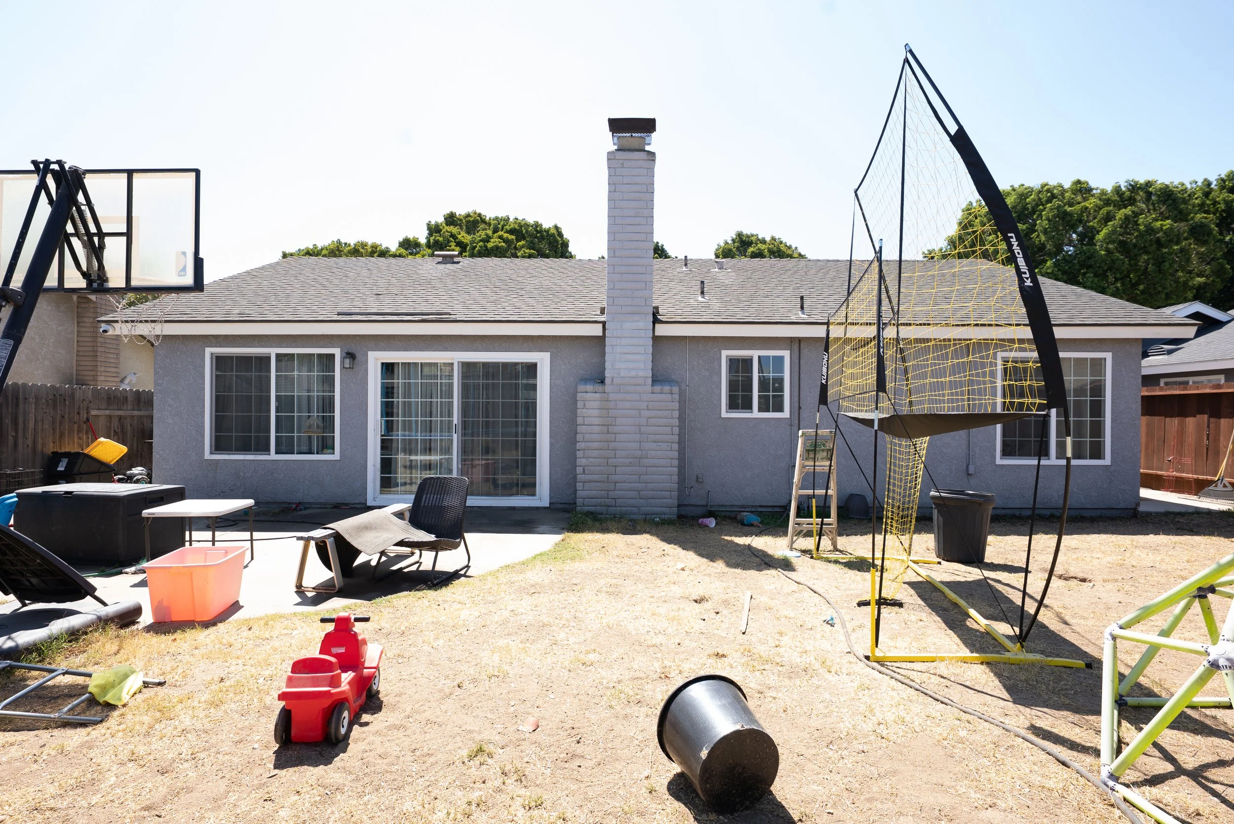 Backyard scene with children's playground equipment, a basketball hoop, scattered toys, and a house with a chimney and patio doors.