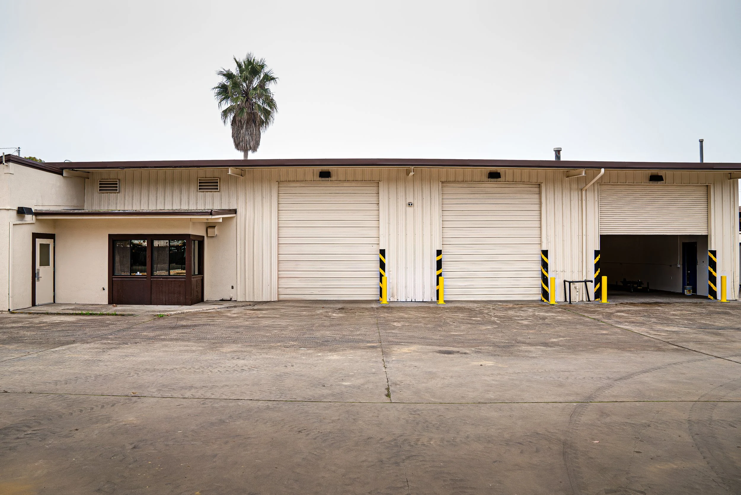 A beige industrial building with three garage doors, one open, and yellow and black safety bollards in front. A tall palm tree is visible behind the building.