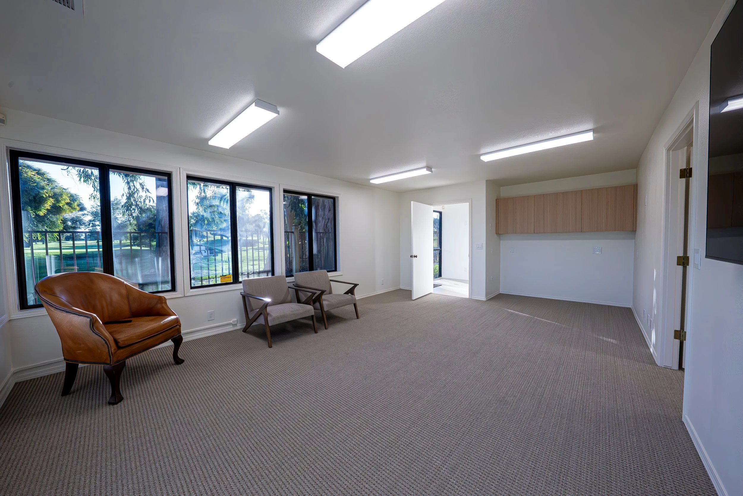 An empty living room with large windows, beige carpet, wooden furniture, and white walls, illuminated by ceiling fluorescent lights.