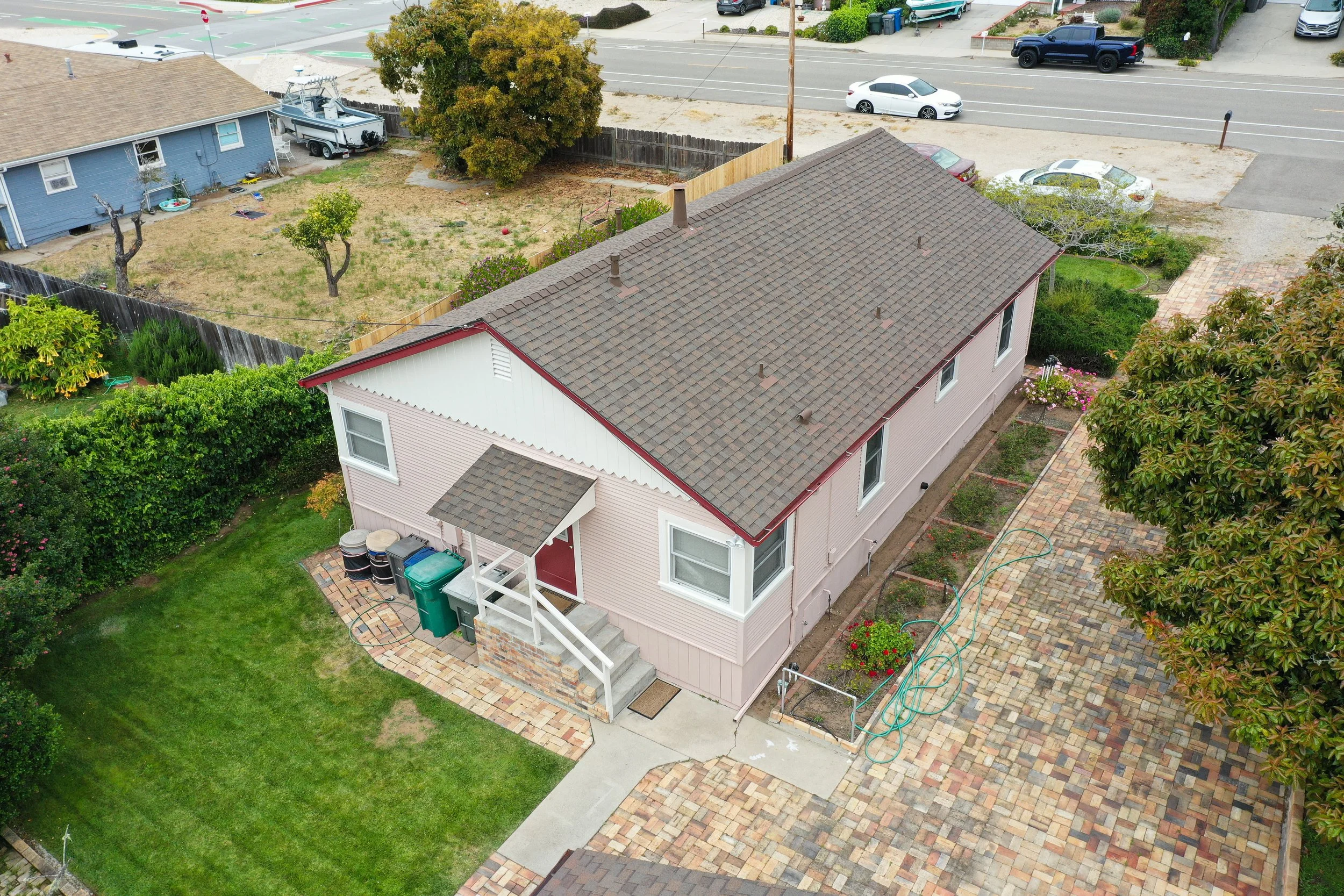 Aerial view of a pink house with a brown shingle roof, surrounded by a lawn, trees, and a brick patio. Multiple trash bins are near the house, and a garden hose is on the patio. The backyard has a small grassy area and some trees, with a tarp-covered