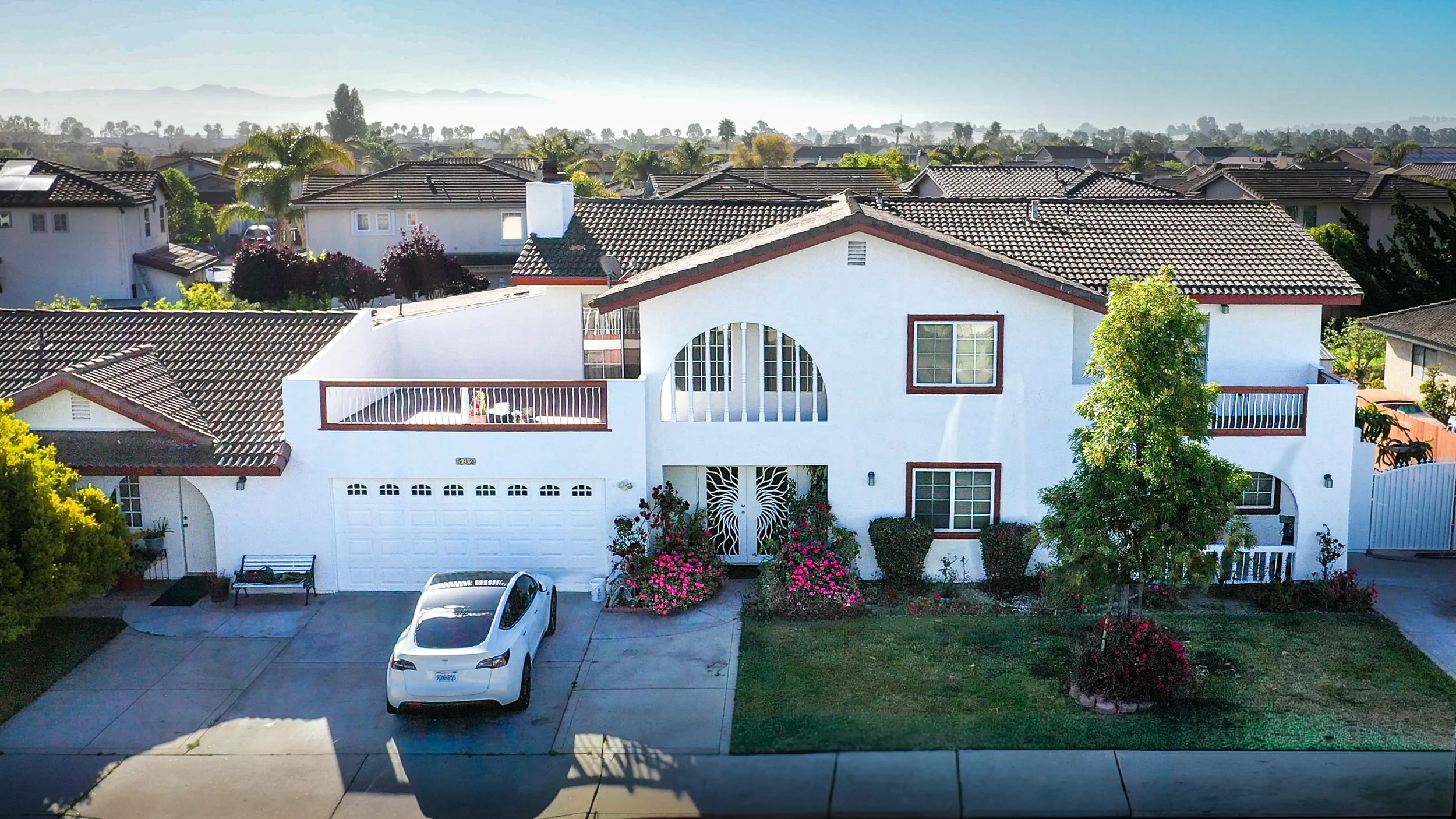 Front view of a white two-story house with a tiled roof, surrounded by greenery and colorful flowers, with a driveway and parked white Tesla in front.