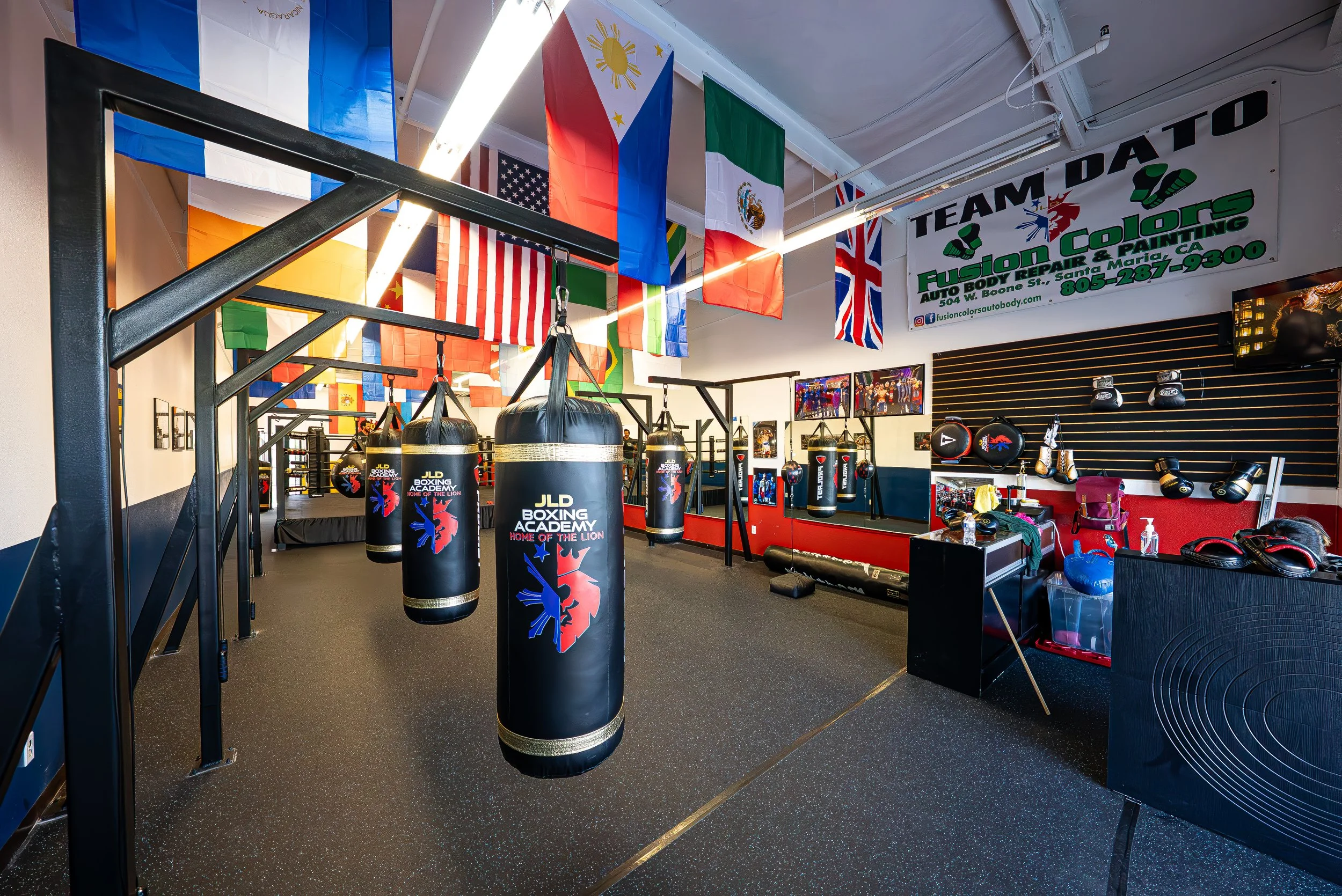 Interior of a boxing gym with punching bags, boxing gloves, and fitness equipment. International flags hang from the ceiling, including flags of the Philippines, United States, Mexico, Italy, and the United Kingdom. A sign for Fusion Colors Auto Body