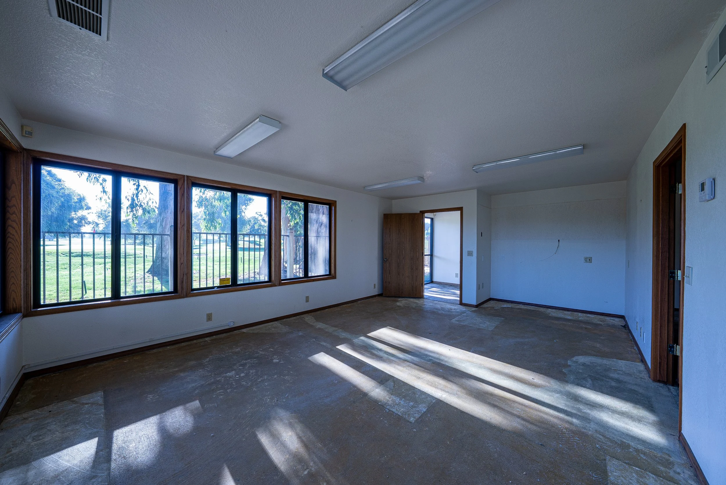 Empty room with large windows, hardwood trim, and bare floor, sunlight casting shadows through windows.