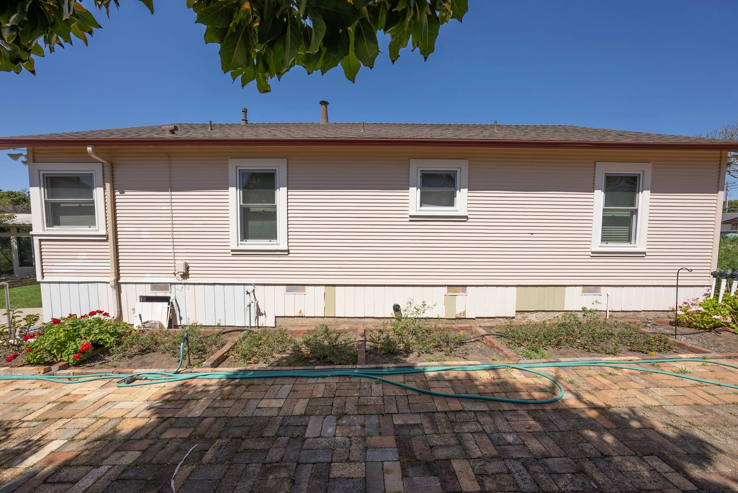 Back of a house with beige vinyl siding and four windows with white trim, a garden with blooming red flowers, a brick patio, and a blue sky with some green leaves from a tree overhead.