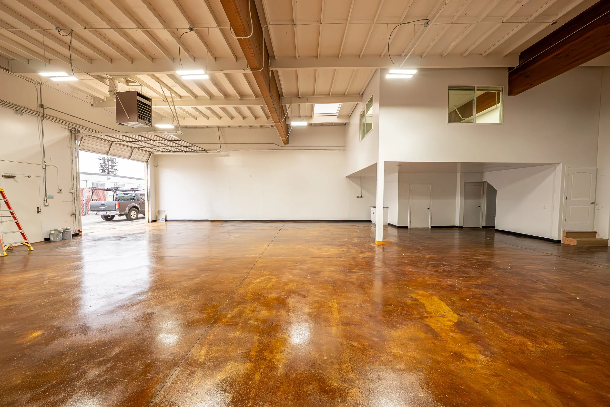 Empty spacious interior with wooden floors, white walls, high ceiling with wooden beams, windows, and overhead lighting. A truck is visible outside through open garage doors.
