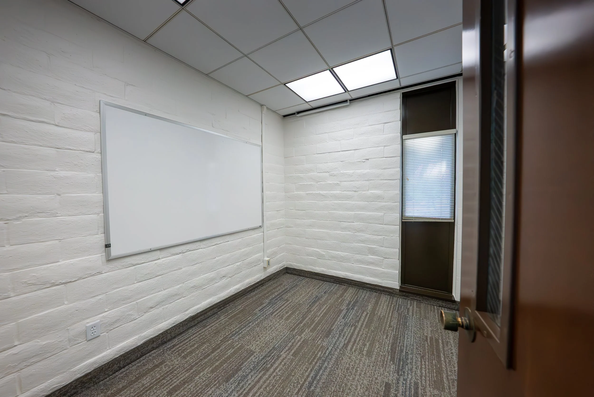 Empty classroom with white brick walls, a whiteboard, a window with blinds, ceiling tiles, and carpeted floor, as viewed from an open door.