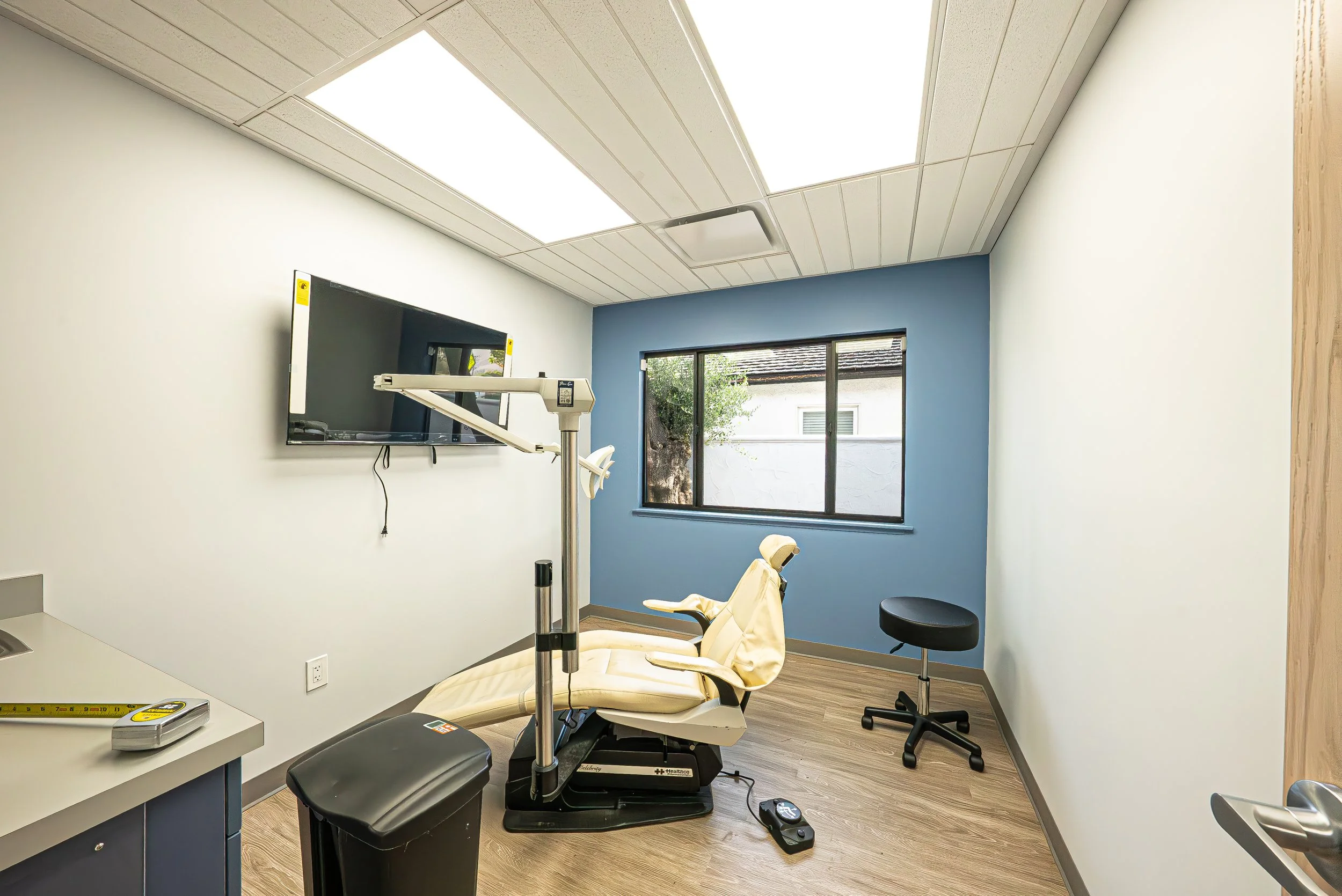 Medical examination room with a dental chair, medical equipment, a mounted television, and a window with a view of the outside.